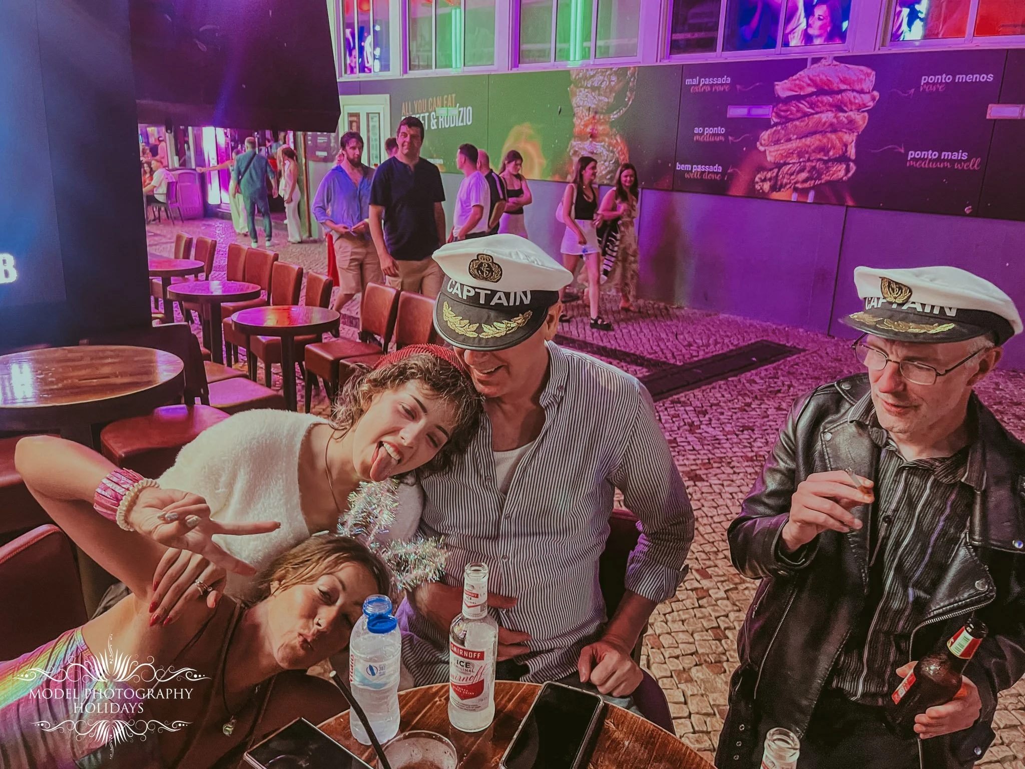 Group of people celebrating at a restaurant or bar, some wearing captain hats, with drinks on the table and colorful neon lights in the background.