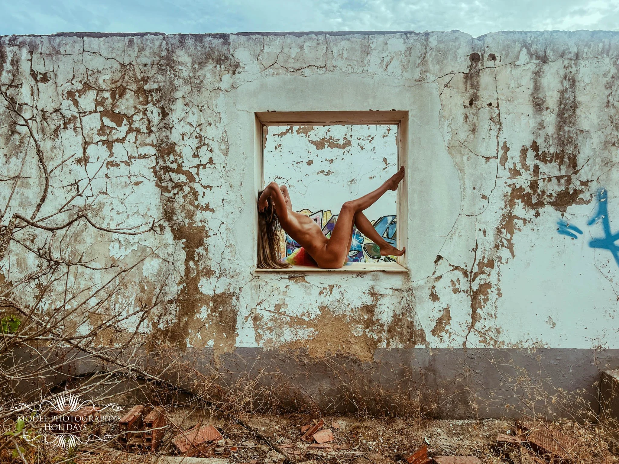 A dilapidated, weathered white wall with cracks and peeling paint, with a rectangular window opening revealing a person lying down, partially covered by a colorful blanket, in front of graffiti on the wall.