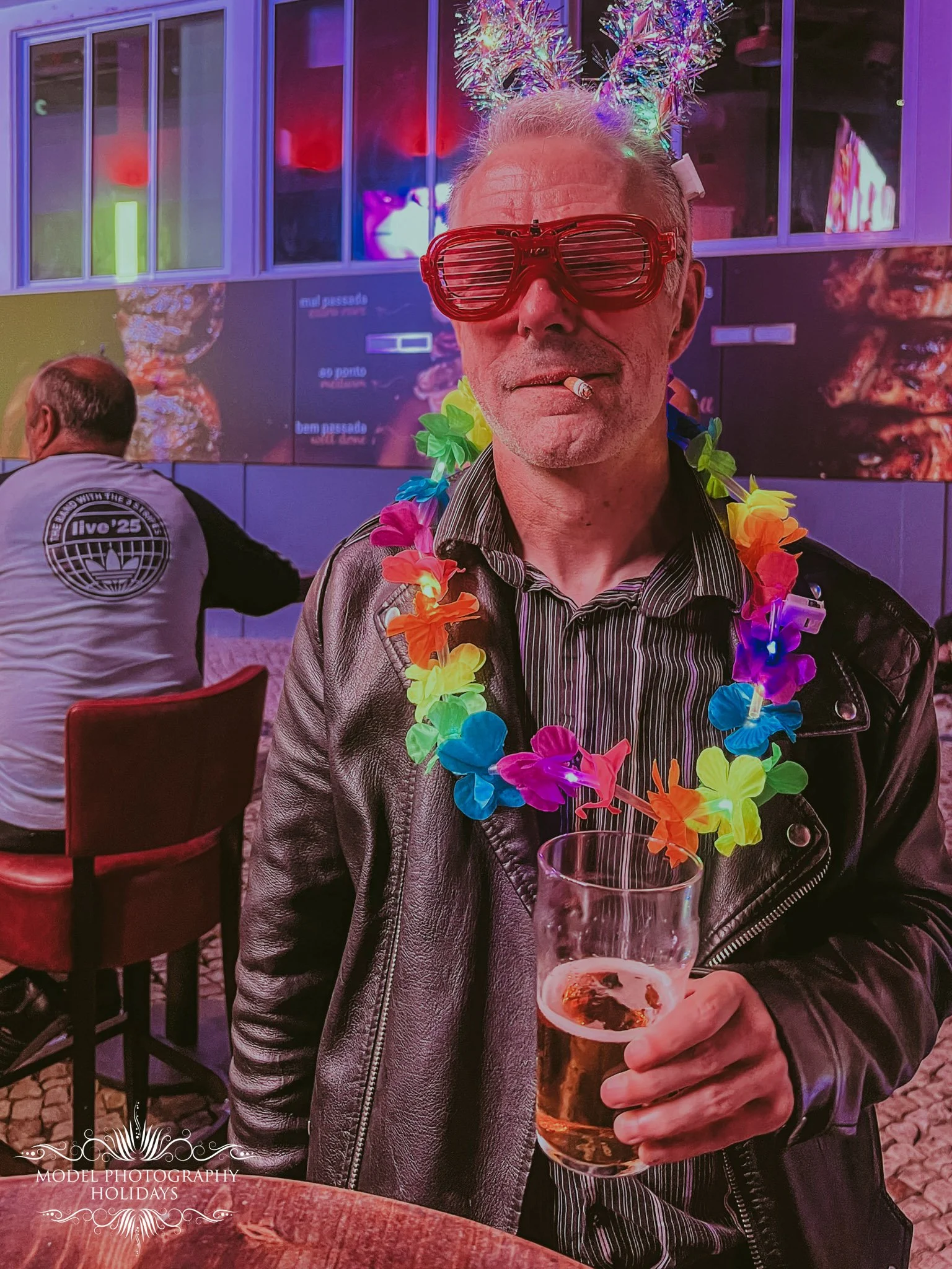 A man with gray hair, wearing red sunglasses, a leather jacket, a colorful lei, and a cigarette in his mouth, holding a glass of beer, at a lively indoor event with a neon-lit background.
