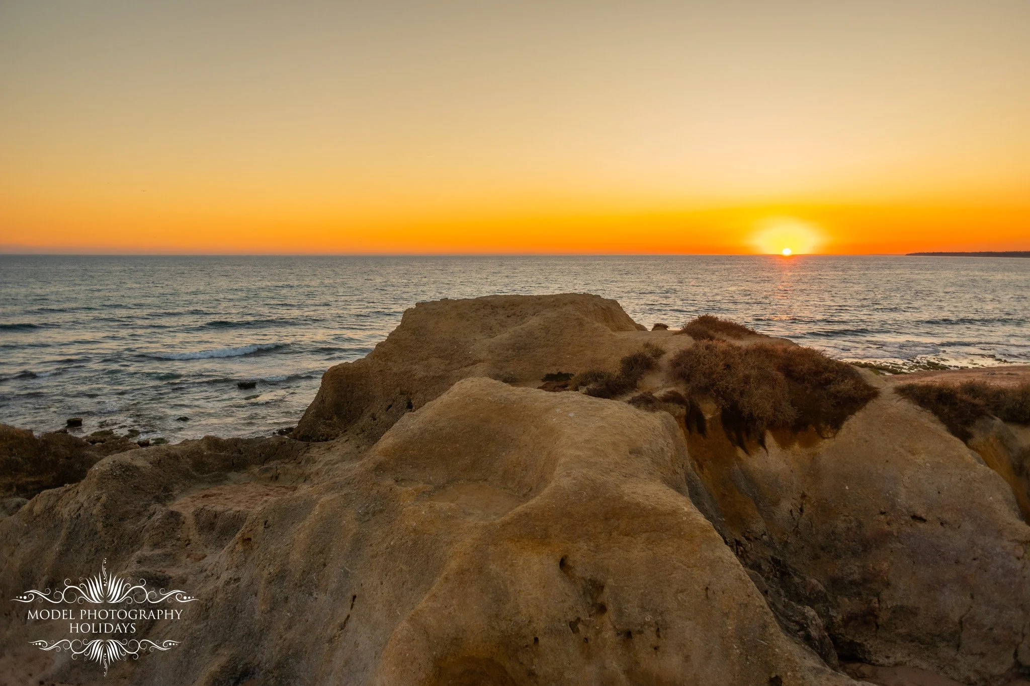 Sunset over the ocean viewed from a rocky shoreline with near rocks in the foreground.