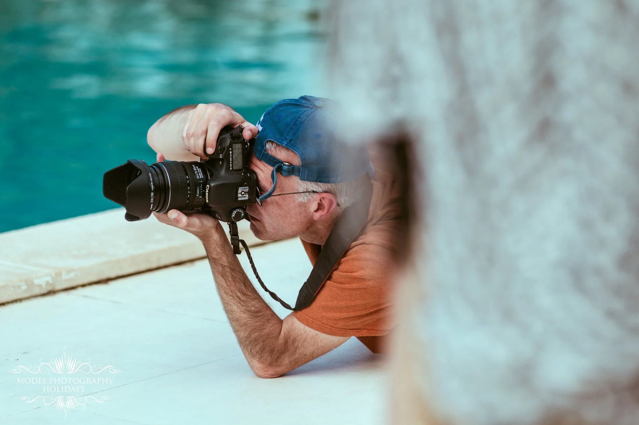 A man with gray hair and glasses, wearing an orange shirt and blue cap, is lying on the ground and taking a photograph with a professional camera.