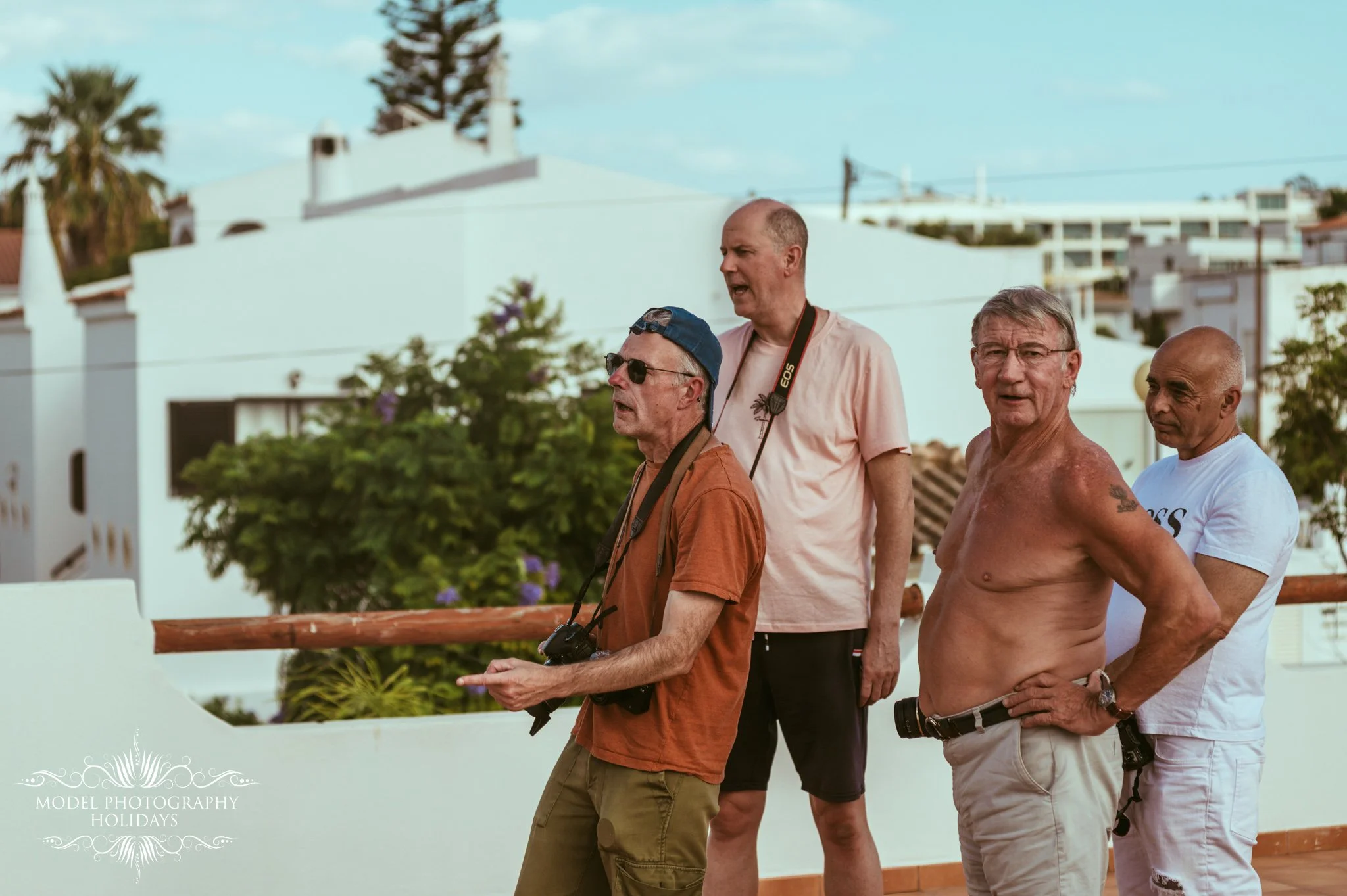 Four middle-aged men walking outdoors near white buildings with trees, one shirtless, others wearing casual clothes, some with cameras, on a sunny day.