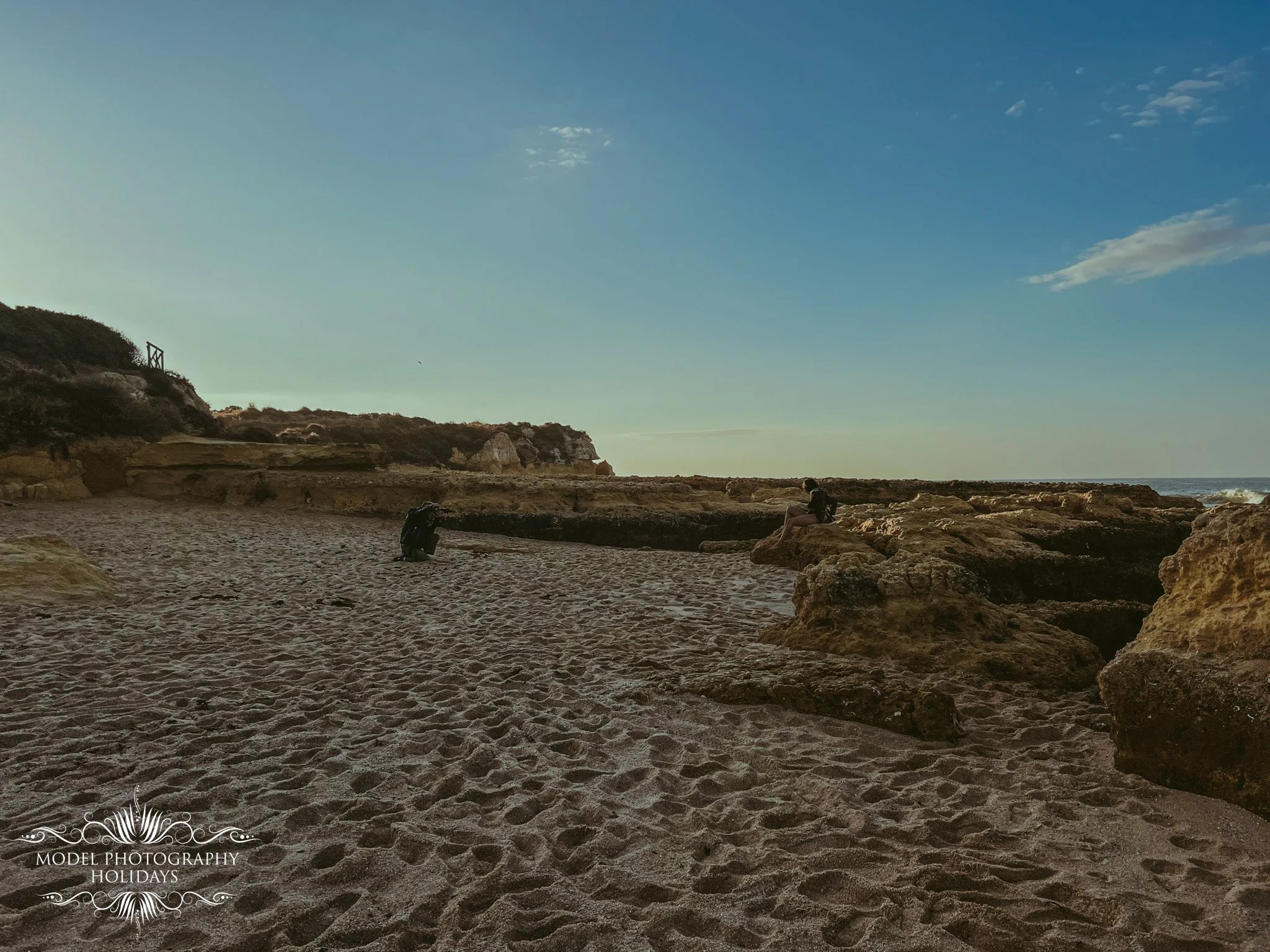 A beach with sandy shore, rocky formations, and a person sitting on a rock near the water, under a clear blue sky with some clouds.