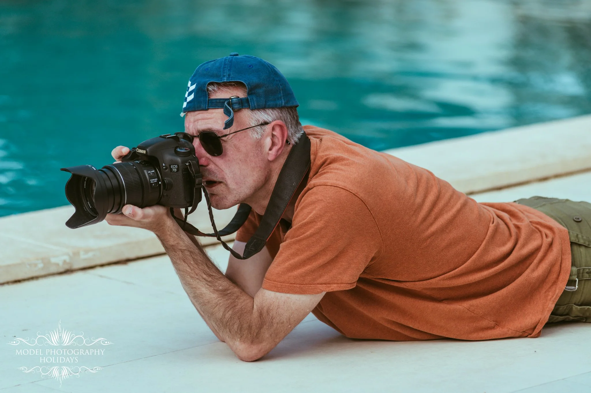 A man lying on his stomach near a body of water, looking through a Canon DSLR camera. He is wearing sunglasses, a blue cap backward, an orange shirt, and olive green pants.
