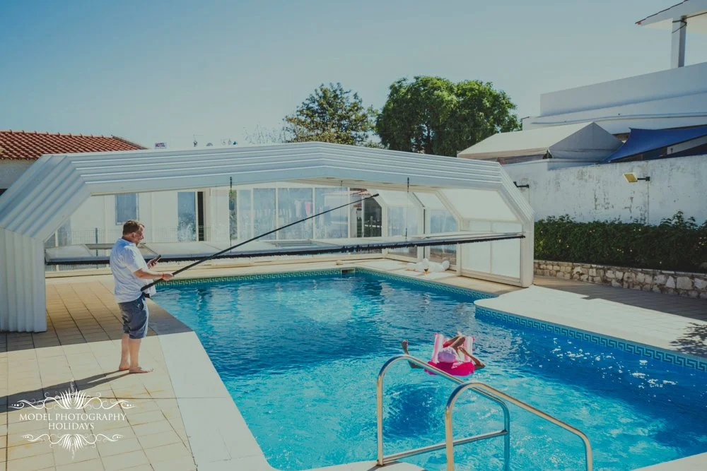 Man cleaning an outdoor pool with a pole near a partially covered area.
