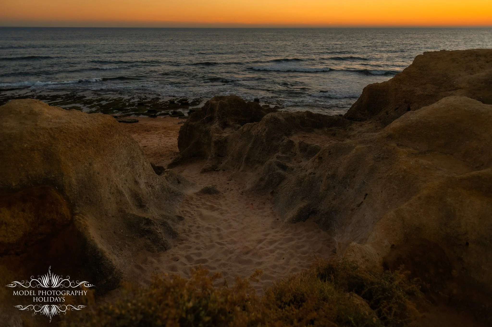 A view from a sandy cove or alcove at sunset, with rocky formations in the foreground and gentle waves on the ocean in the background. The sky is orange and yellow near the horizon.
