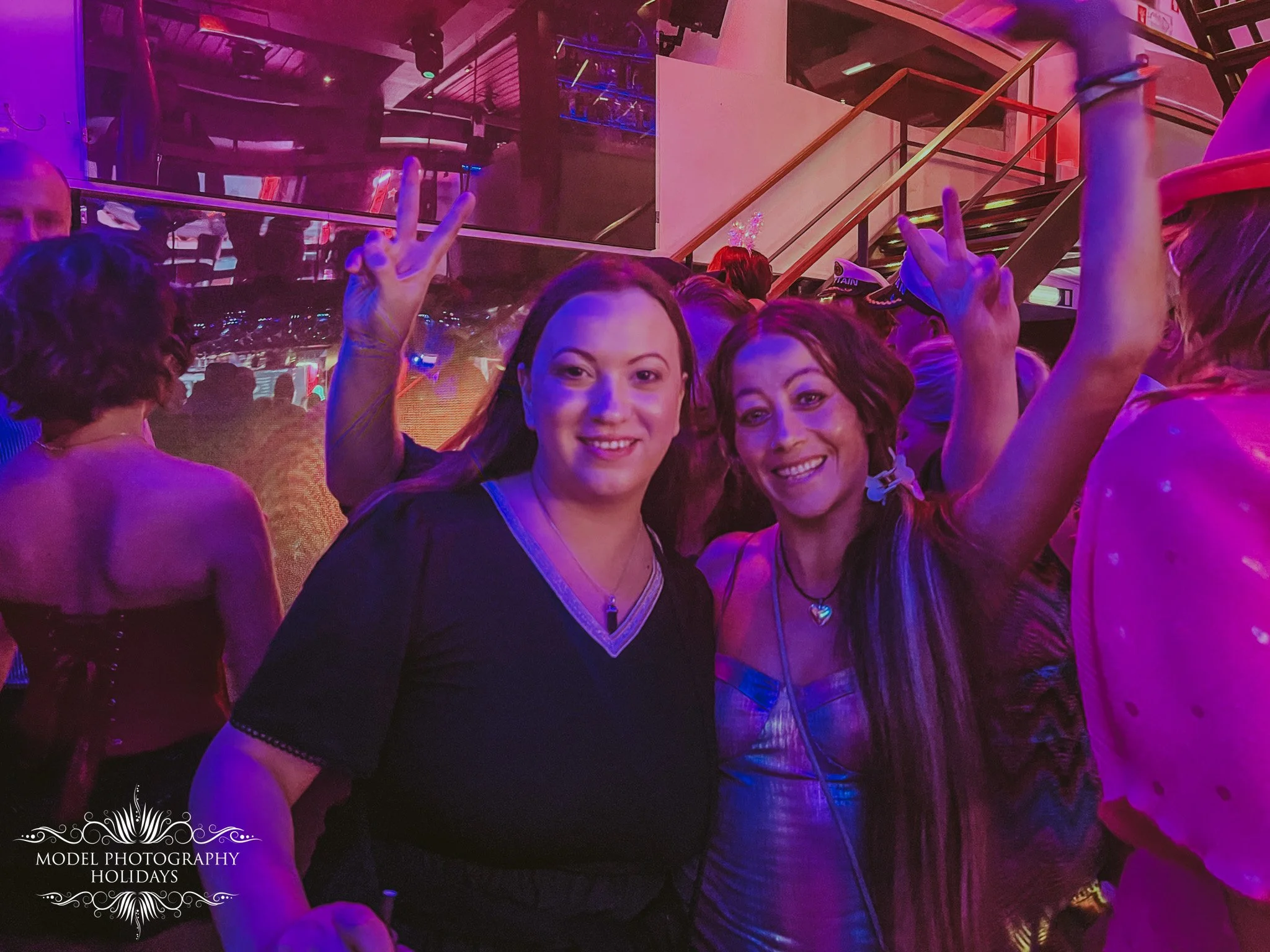 Two women smiling and making peace signs at a party or nightclub with purple and pink lighting, surrounded by other people. One woman is wearing a black dress with a heart-shaped necklace and the other has long hair with a white hair accessory.