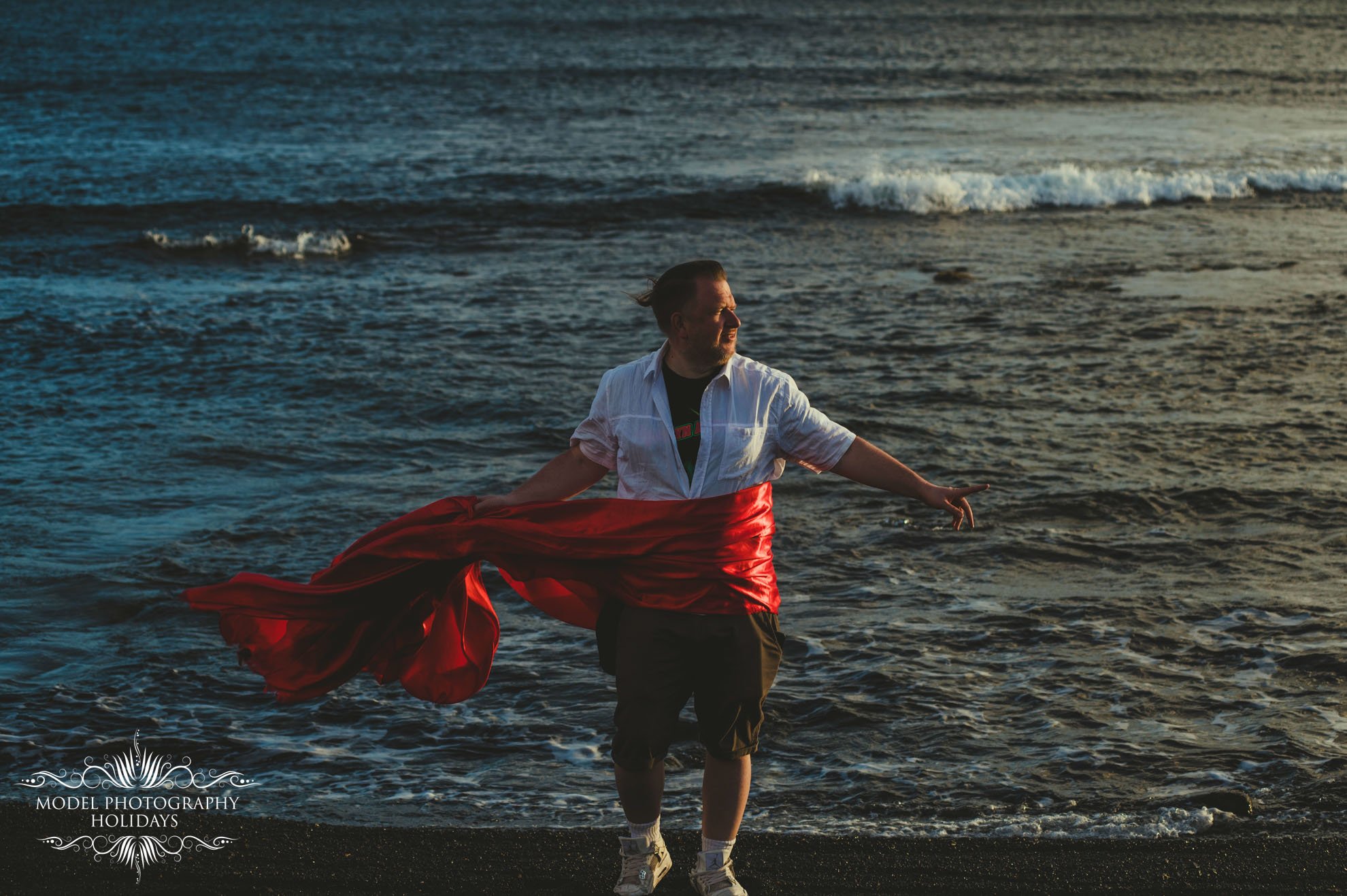 Person standing on a beach with waves, wearing a white shirt and wrapped in a red fabric.