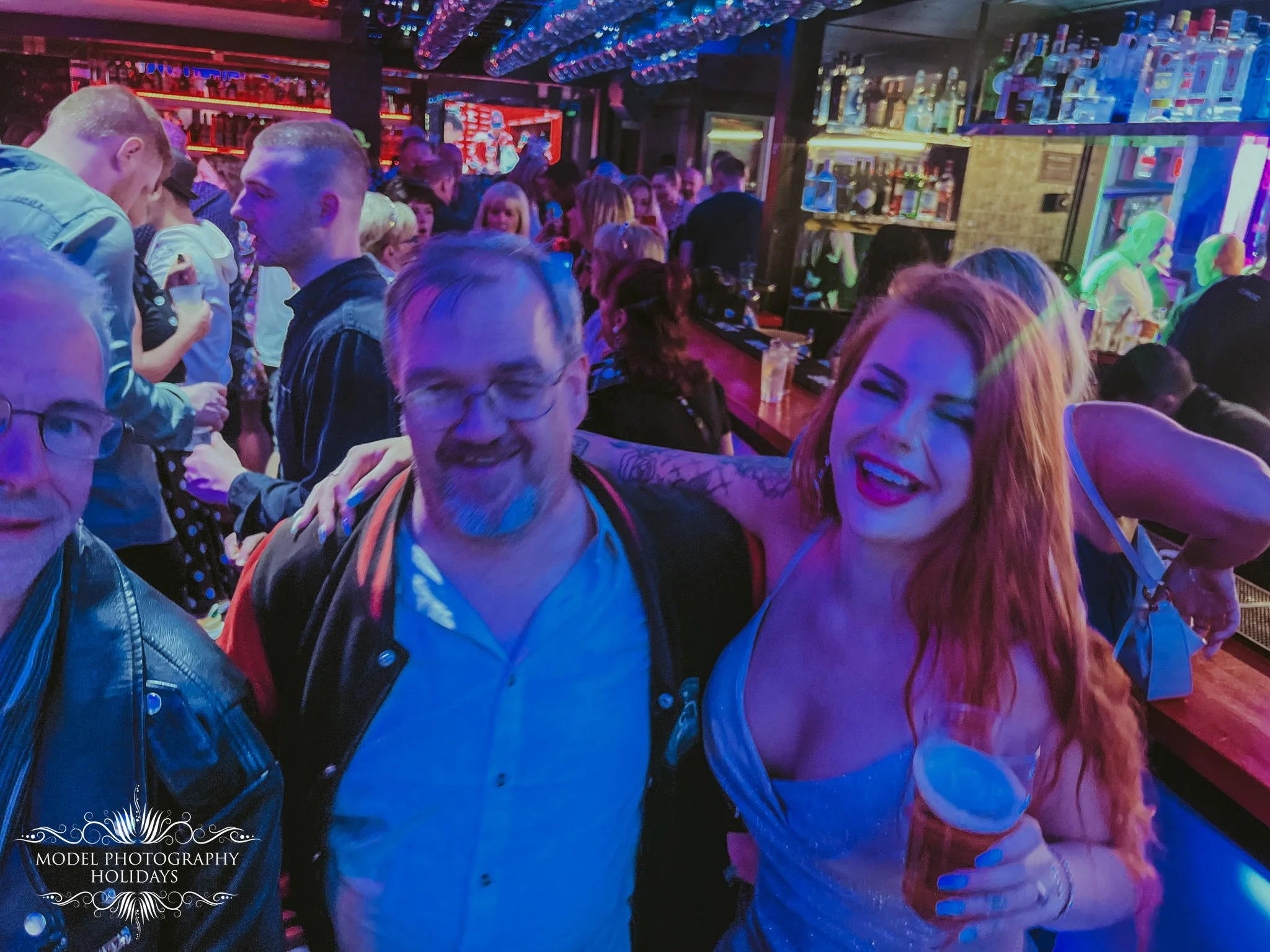 People socializing in a dimly-lit bar with colorful neon lights, shelves of liquor bottles, and a lively atmosphere.