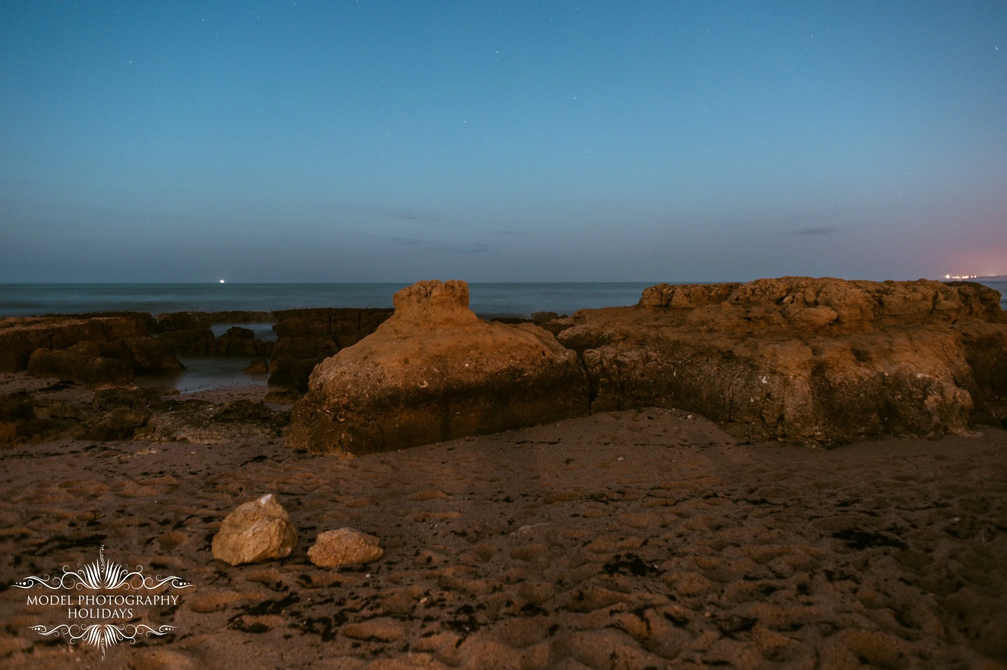 A rocky beach at dusk with large rocks in the foreground, calm ocean water, and a clear sky with little cloud cover.