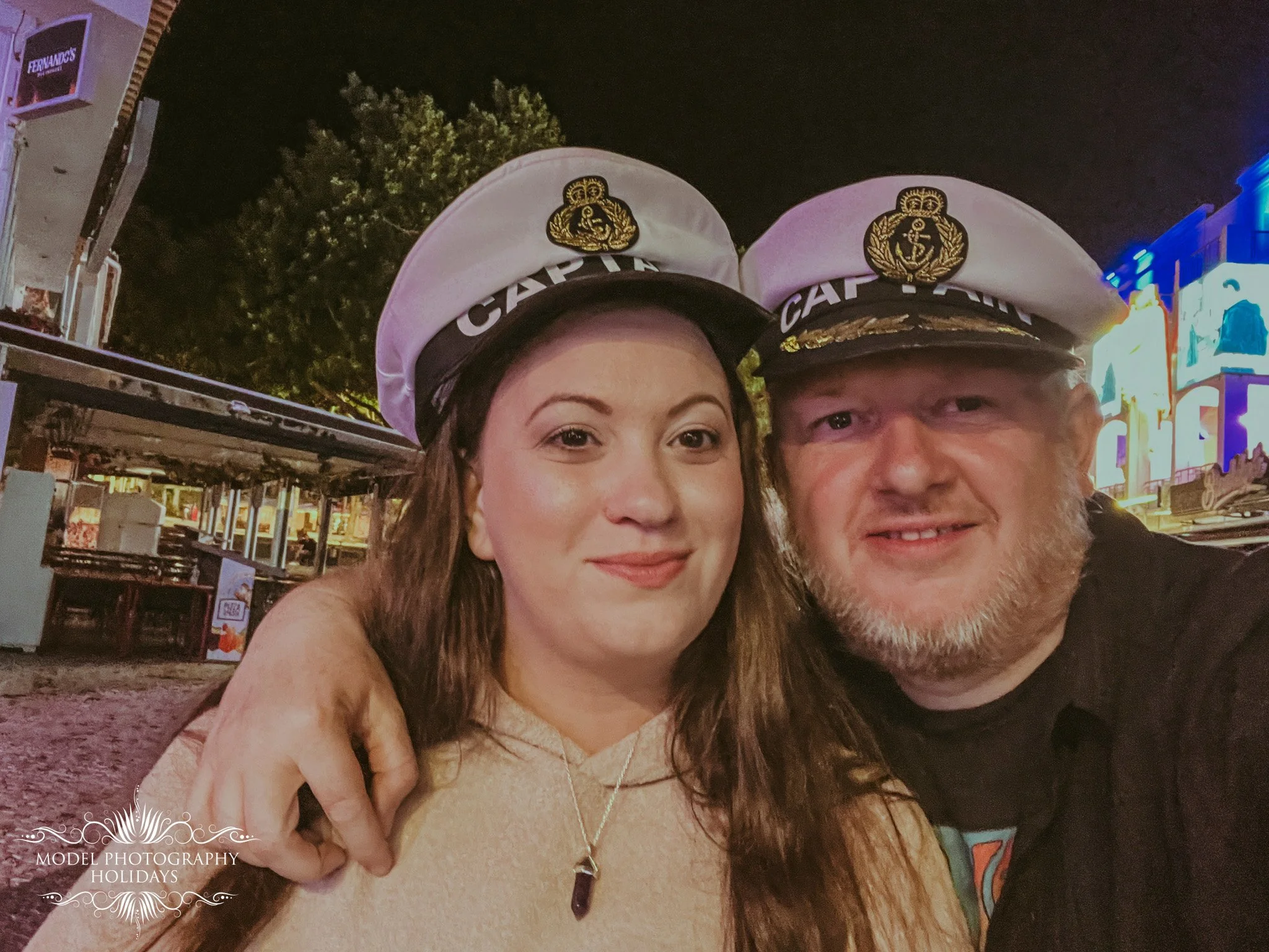 Two people wearing sailor hats with gold badges, taking a selfie outdoors at night, with a background of a small building with a neon sign and trees.