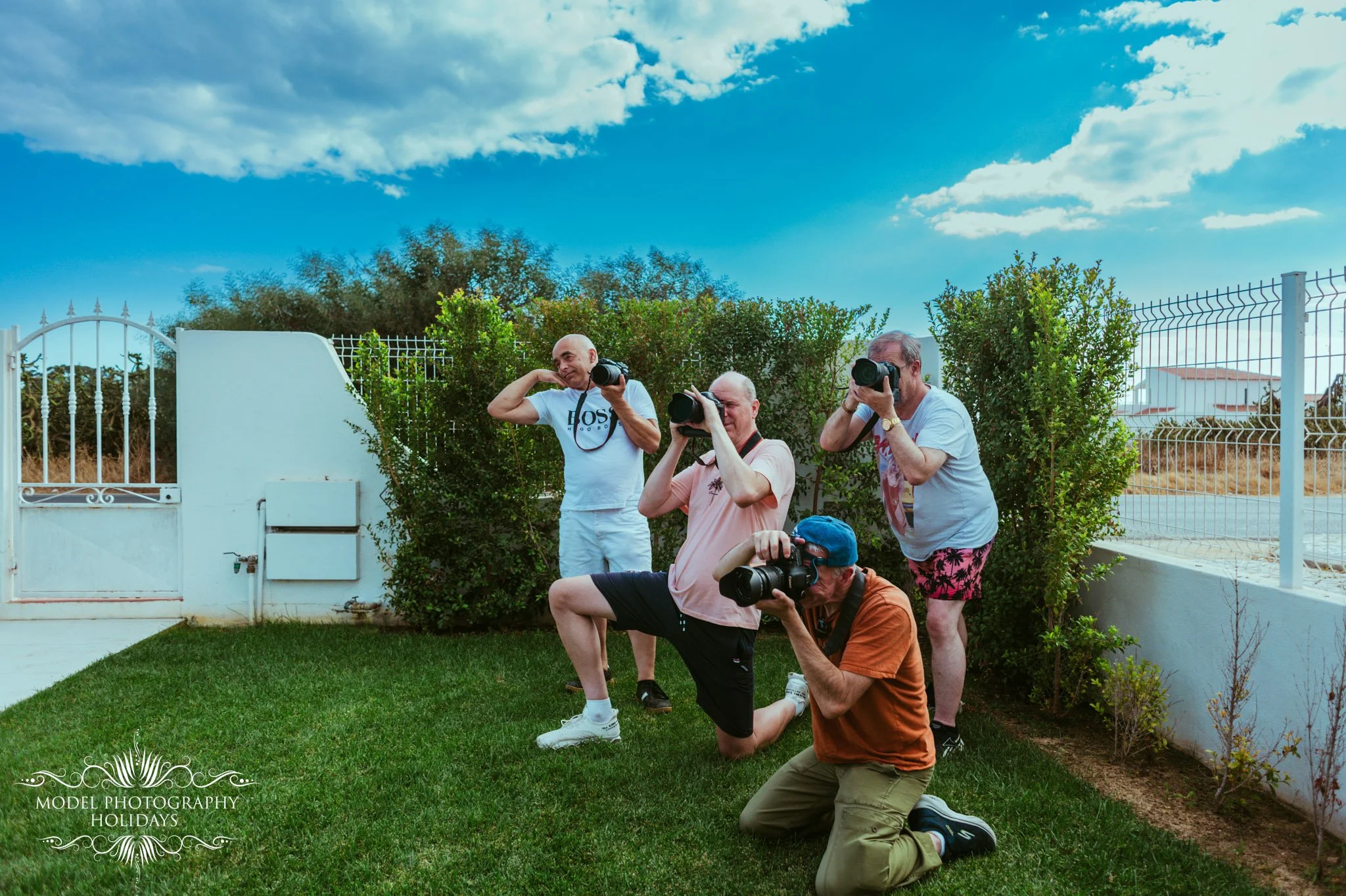 A group of five men, some standing and some kneeling, taking photos outdoors. They are in a backyard with a white fence, greenery, and a bright blue sky with clouds. The men are holding cameras and posing in different positions.