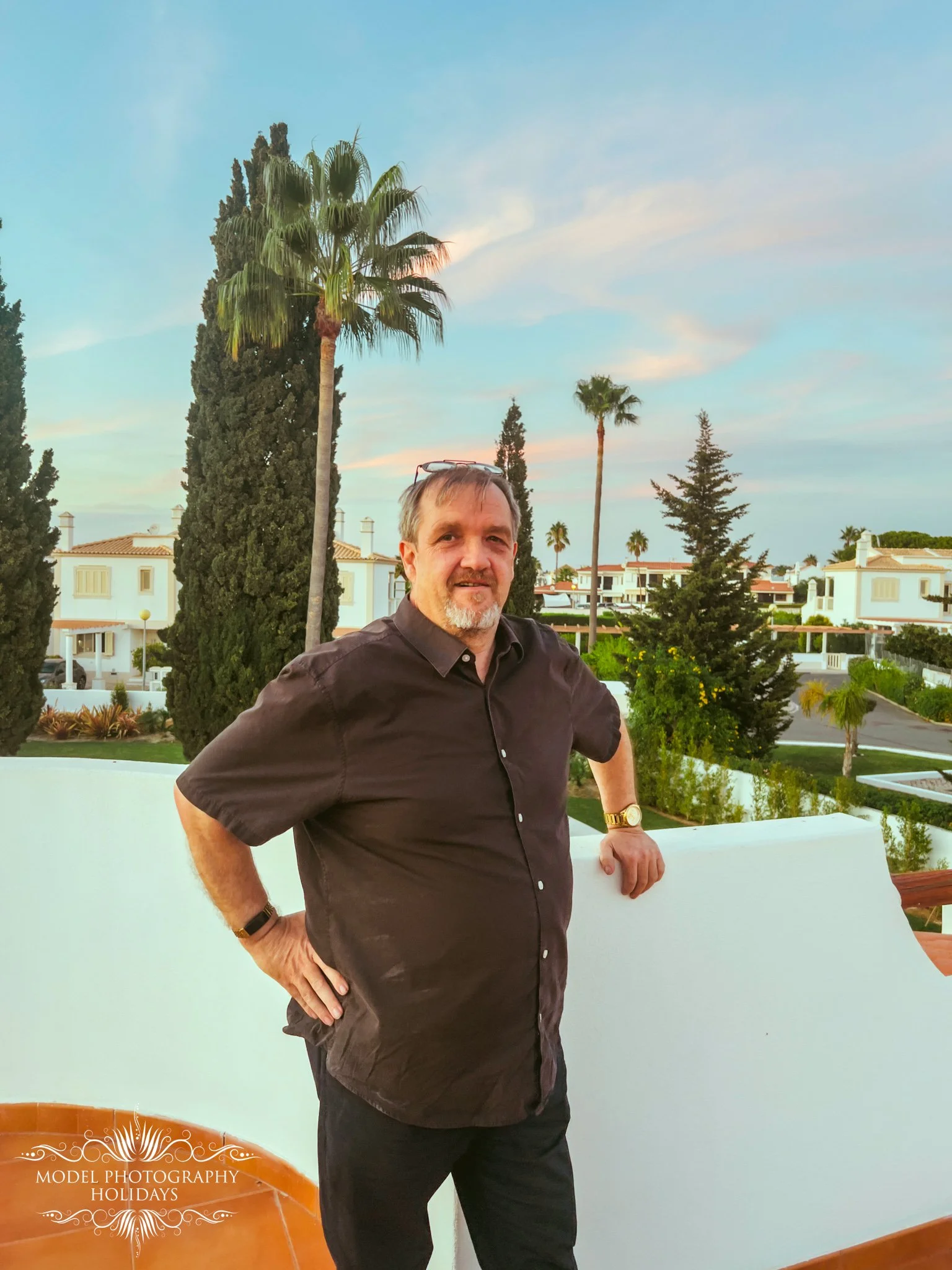 A man in a dark shirt with glasses on his head and a gold watch stands on a balcony with white walls, surrounded by tall palm trees and lush greenery, with a background of residential buildings under a blue sky with some clouds.