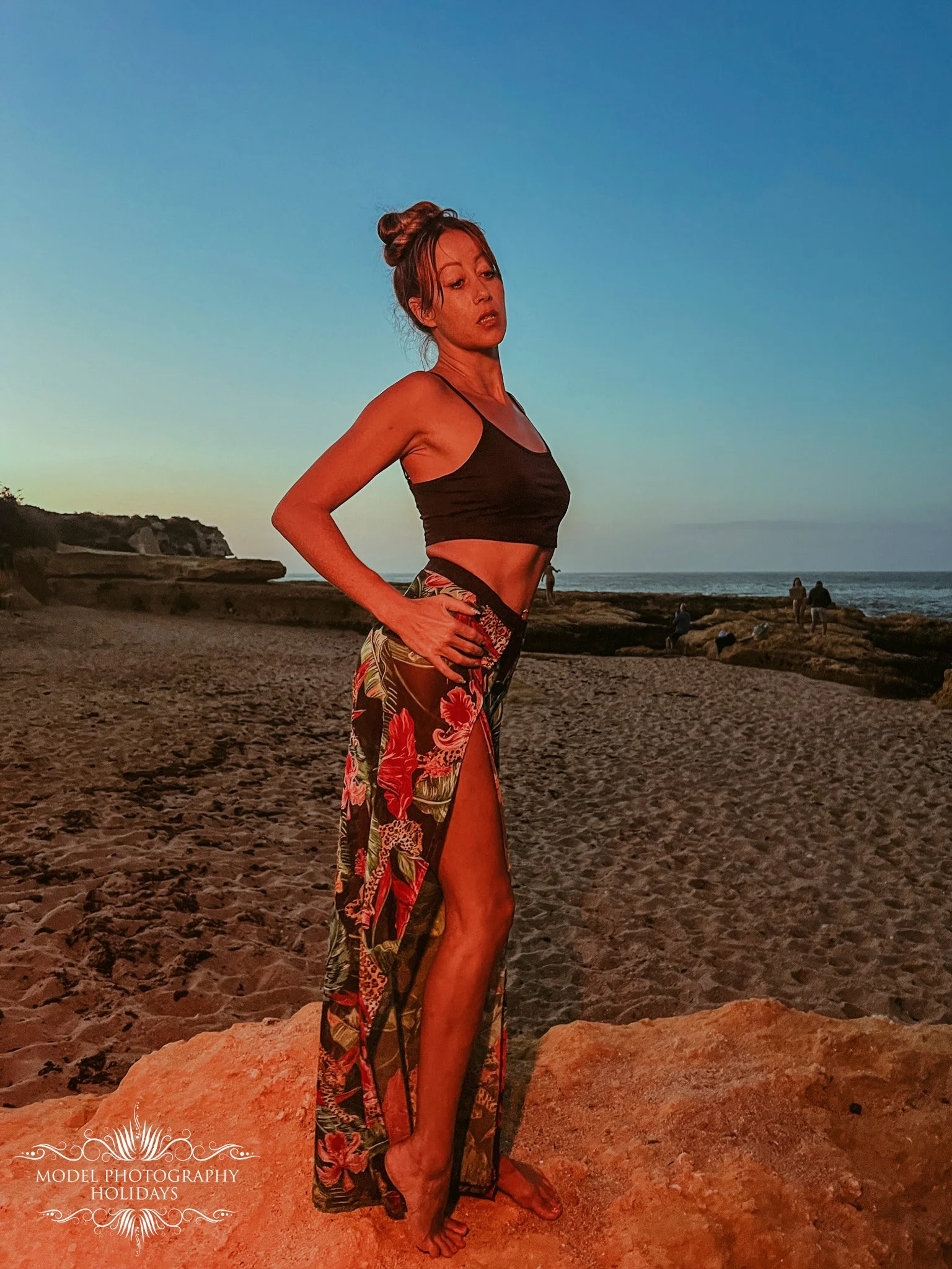A woman is standing on a sandy beach with rocks, wearing a black crop top and colorful floral long skirt with high slit, during sunset or sunrise. The sky is clear and blue, with a few people sitting and standing near the shoreline in the distance.