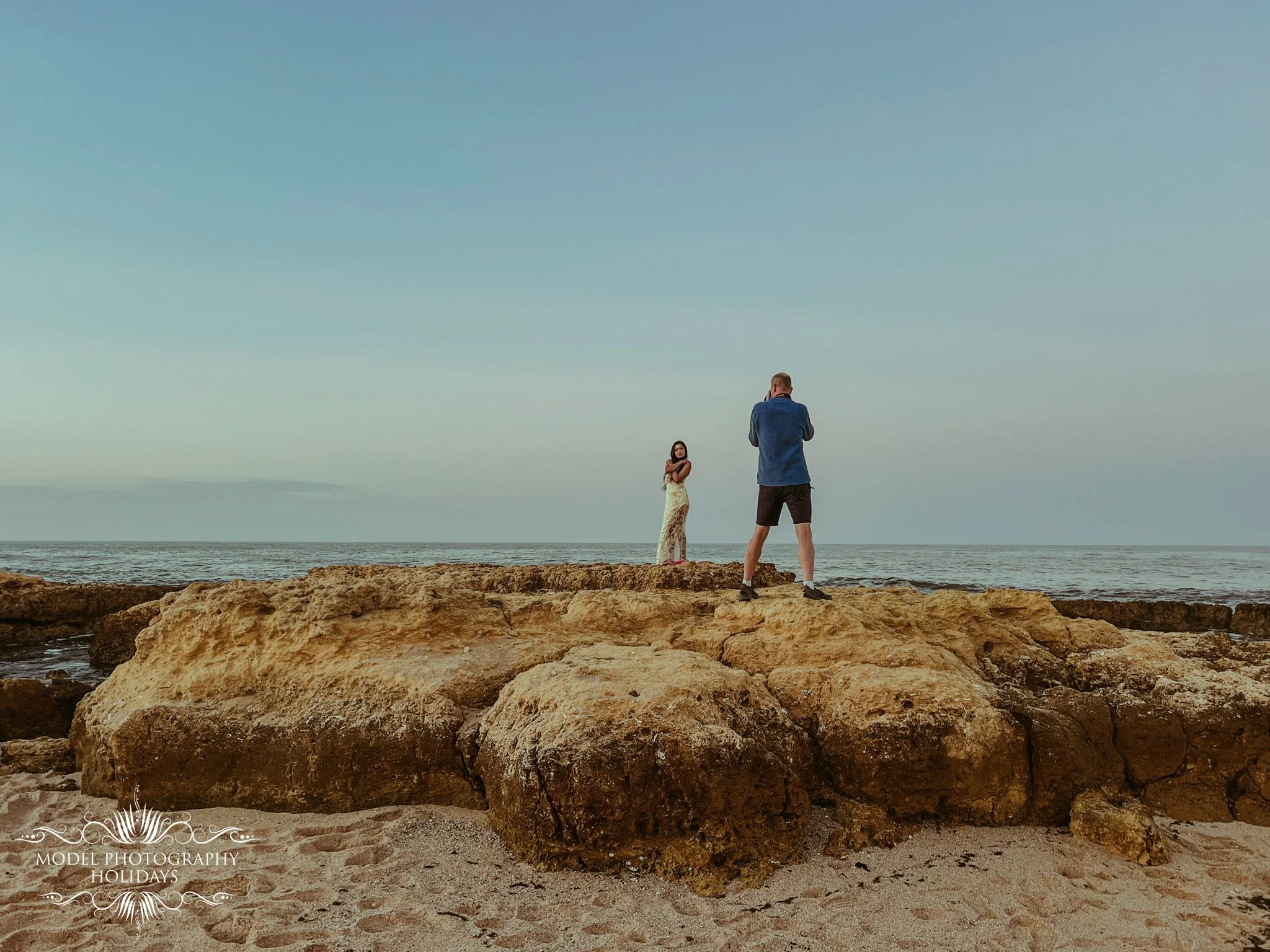A man and a woman standing on rocks near the beach. The man is taking a photo or video of the woman, who is standing with her arms crossed. The sky is clear with minimal clouds, and the scene is peaceful.