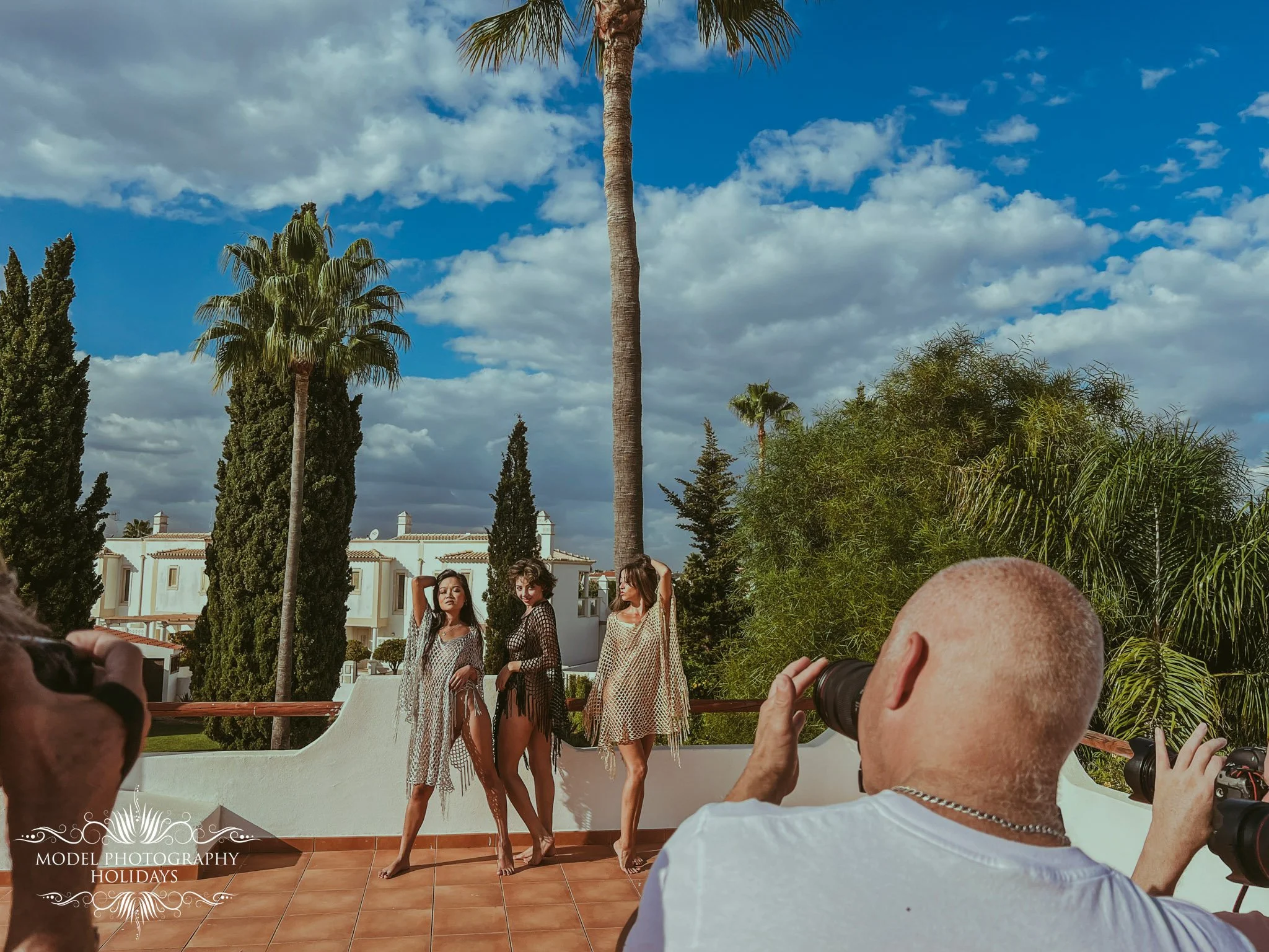 Three women are posing for photos outdoors with a background of palm trees, green foliage, and a clear sky with scattered clouds. Two men are taking pictures of the women, and at least one man is holding a camera. The setting appears to be a sunny da