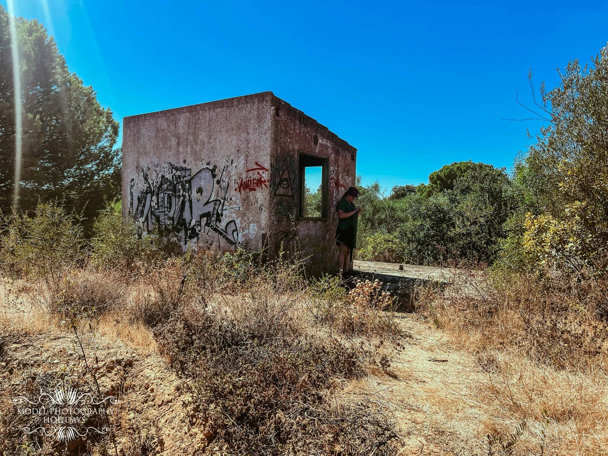 An abandoned building with graffiti walls, surrounded by dry bushes and tall trees under a clear blue sky. A person is standing next to the building, looking at their phone.