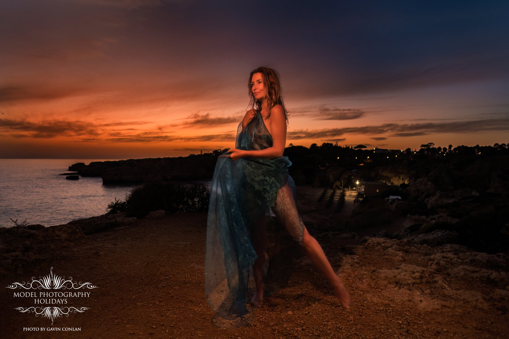 Woman standing on a rocky cliff at sunset, wrapped in a translucent blue fabric, overlooking the ocean.