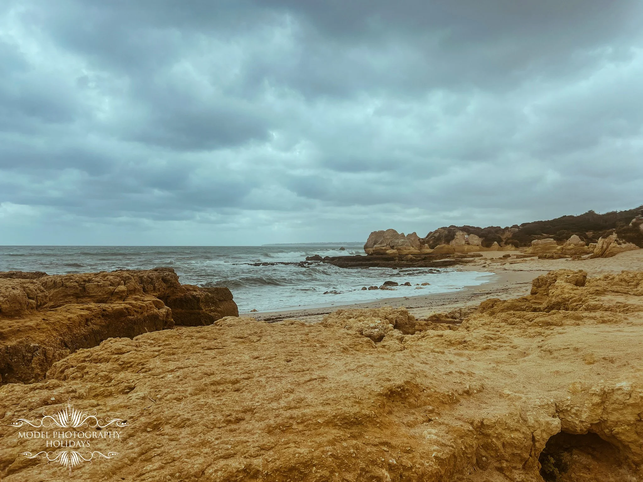 A rocky beach with sand, large rocks, and cliffs in the background under a cloudy sky.