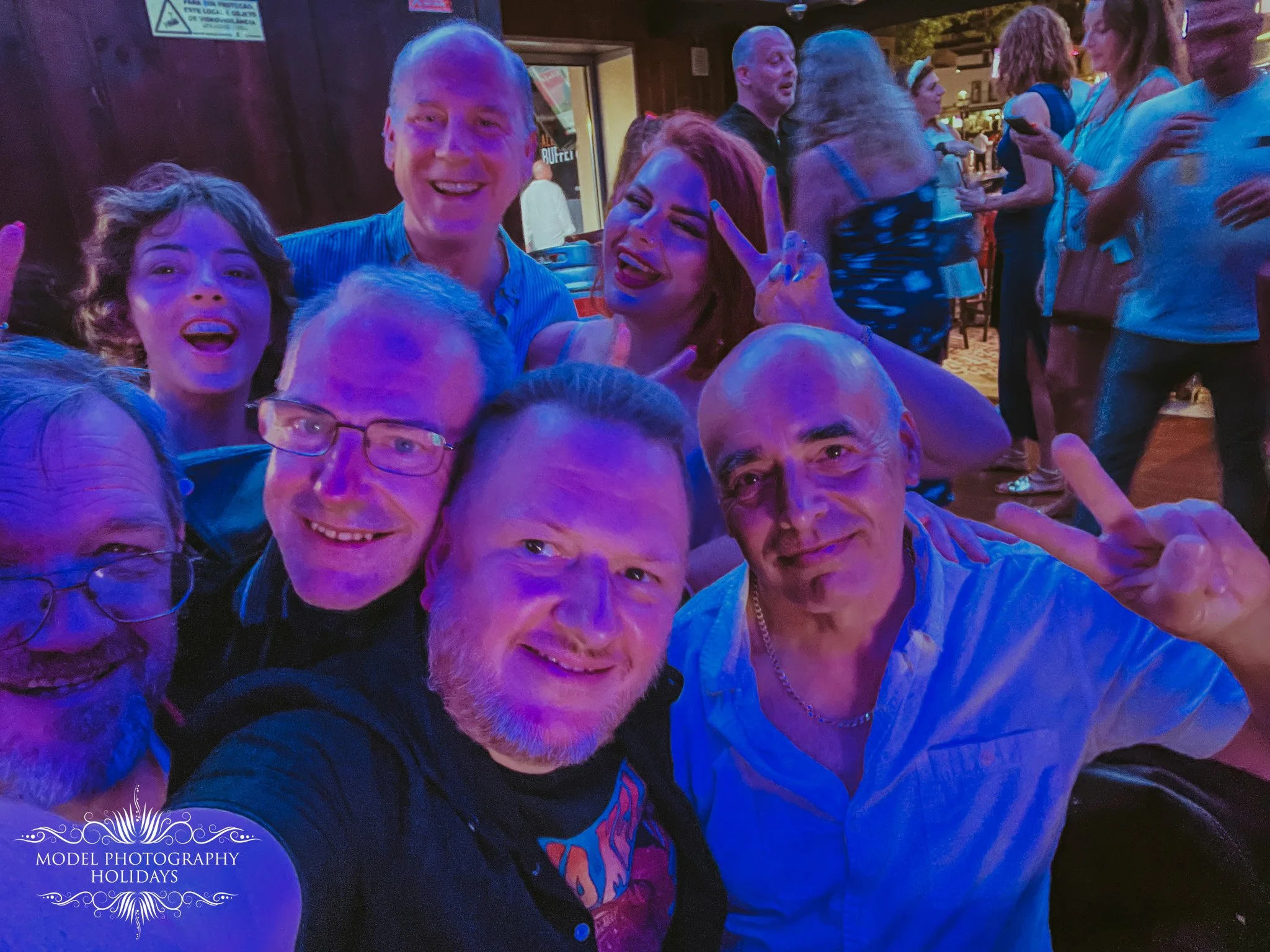 Group of people at a social gathering, smiling and celebrating. One woman is making a peace sign, others are enjoying themselves. The setting appears lively with colorful lighting.