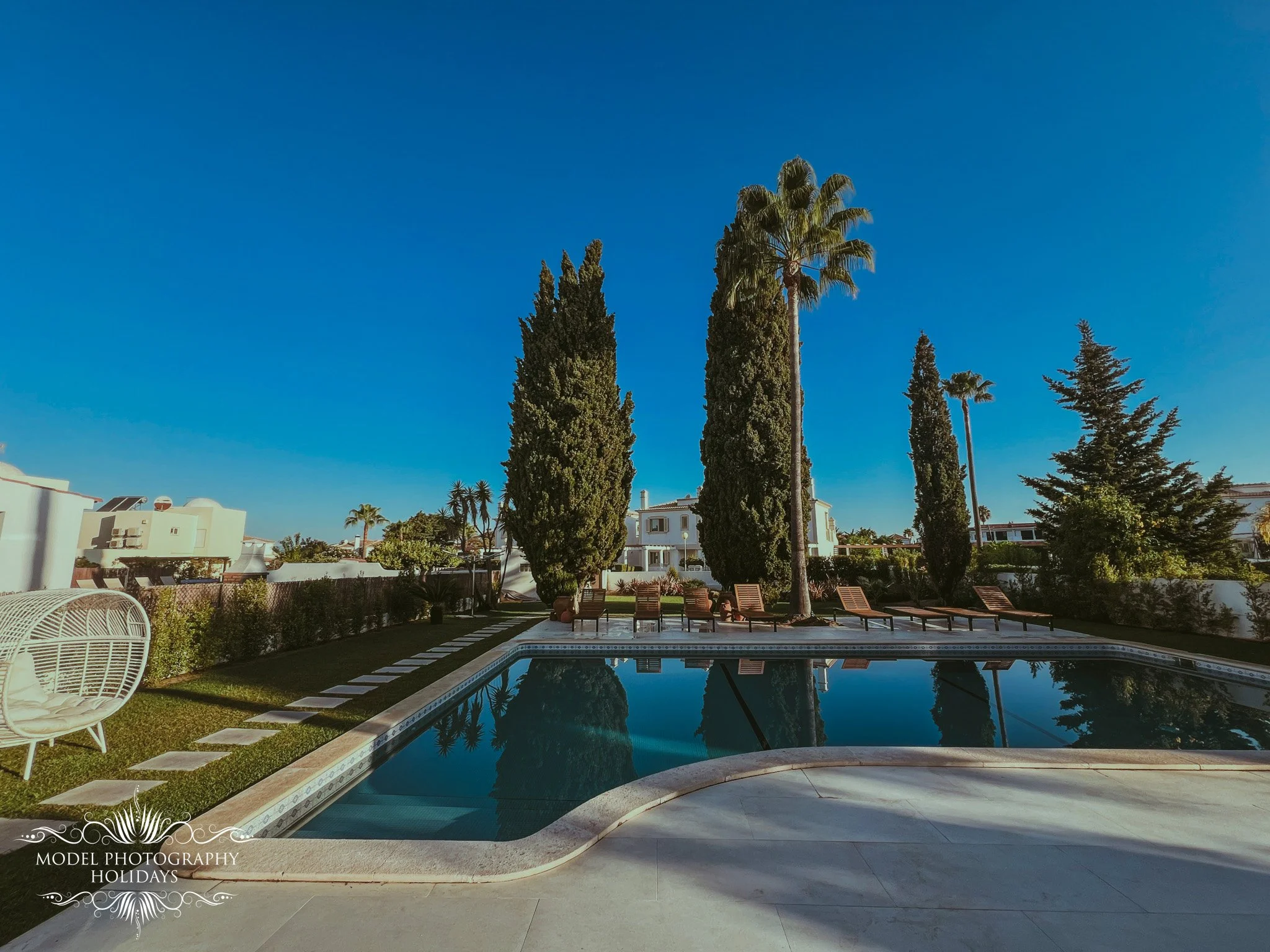A backyard with a swimming pool, surrounded by chairs, palm trees, and other greenery under a clear blue sky.