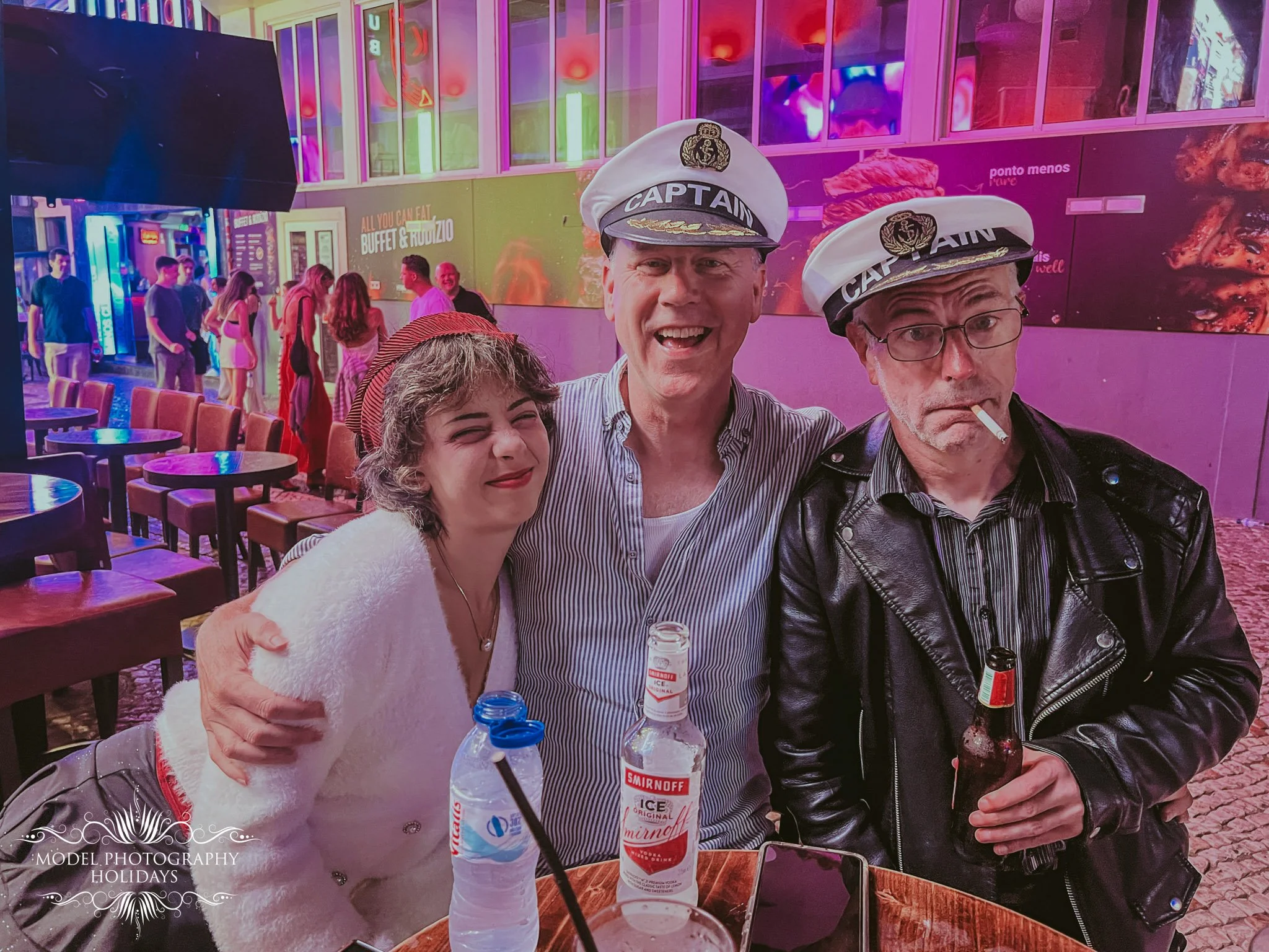 Four people standing together at a bar or club, smiling and wearing captain hats. The background features colorful neon lights and a bar with drinks, with a crowd of people in the background.