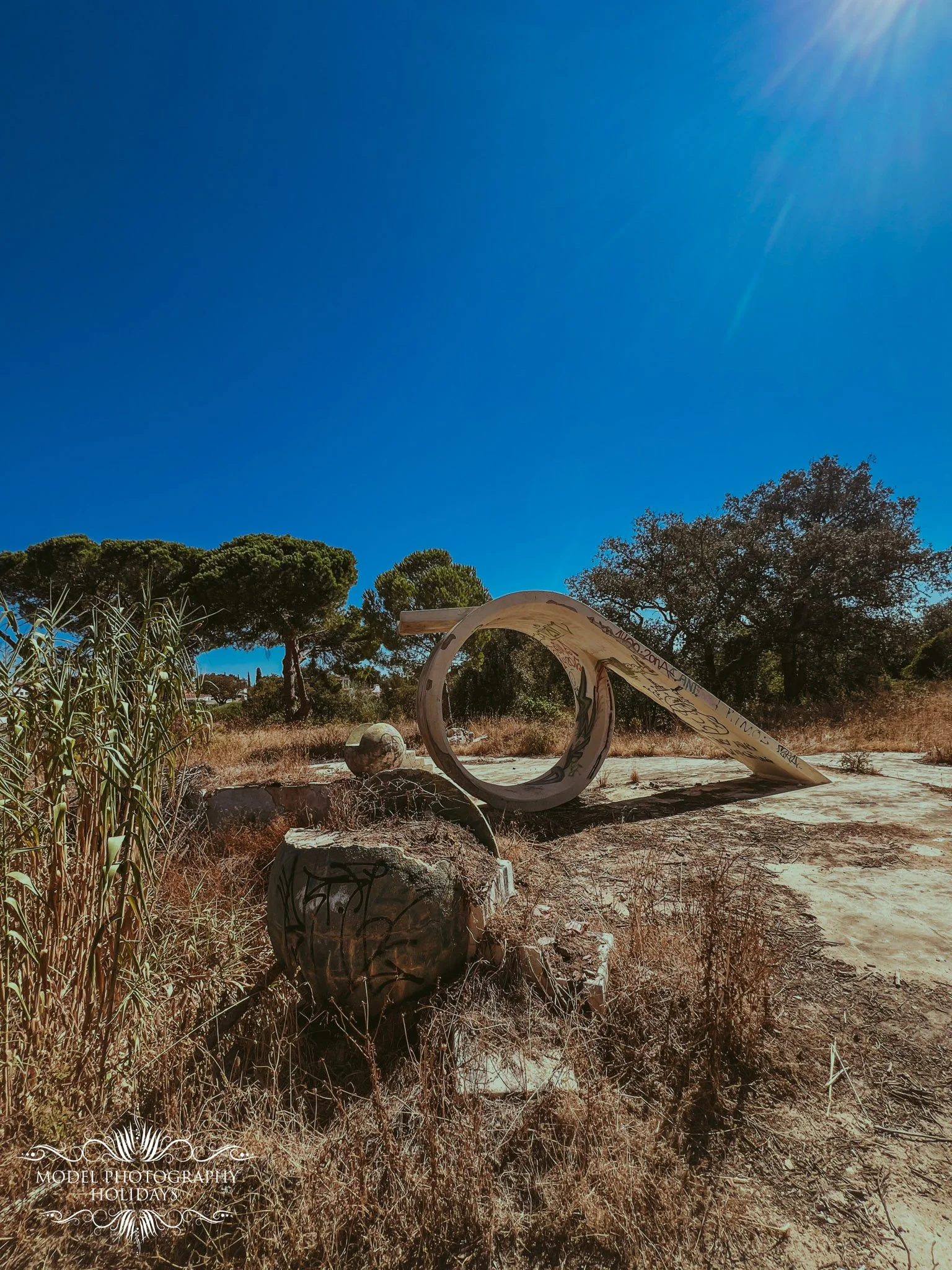 Outdoor scene with dry grass, trees, a concrete sculpture resembling a hoop and a ball, and graffiti on the sculpture, under a clear blue sky with sunlight.