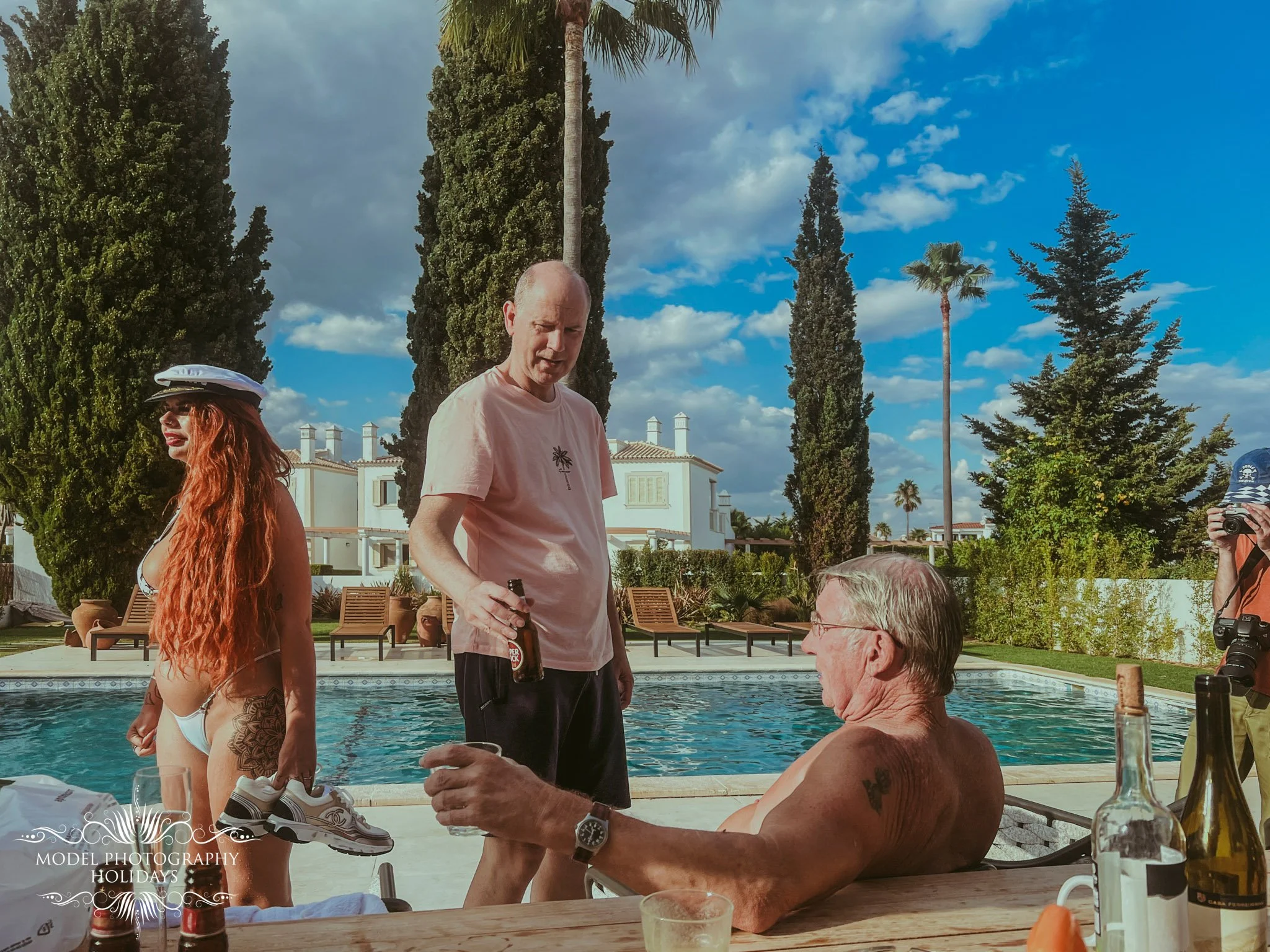 A group of people gathered outdoors around a swimming pool on a sunny day, with trees and a white building in the background. One person is standing, holding a drink, and others are sitting or standing, engaged in conversation.