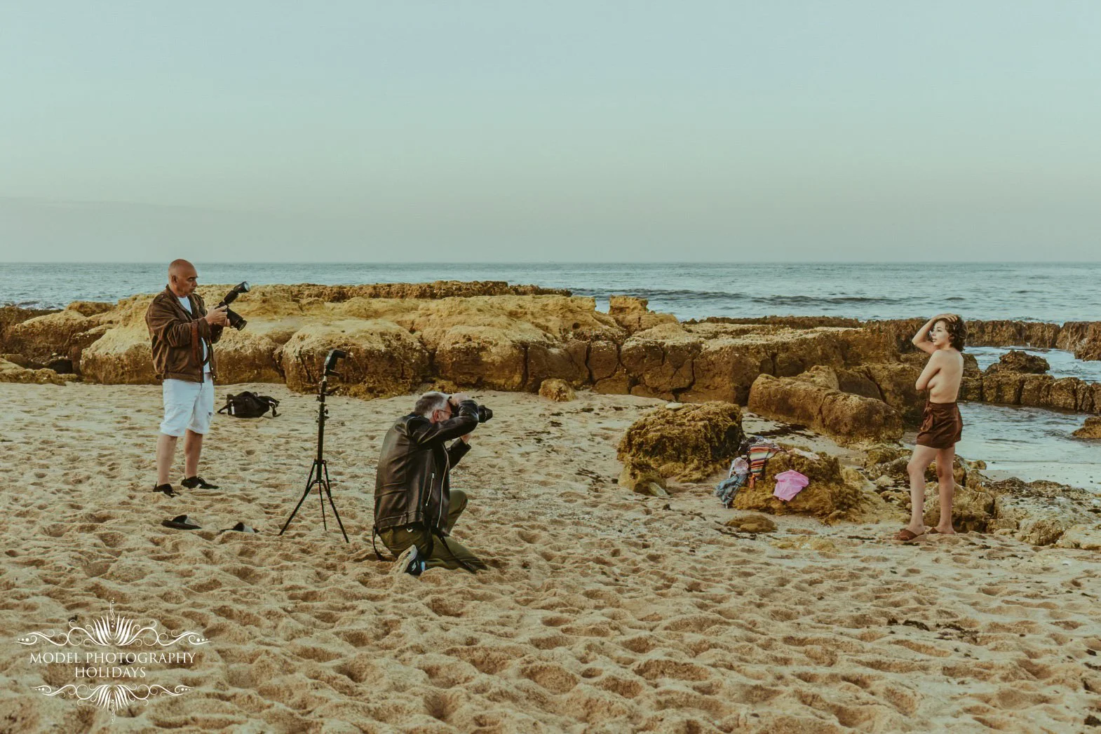 A photoshoot at the beach with a woman posing near rocks while two photographers take pictures of her. One photographer is using a camera mounted on a tripod, the other is kneeling with a camera, and a man observes holding another camera. The beach h