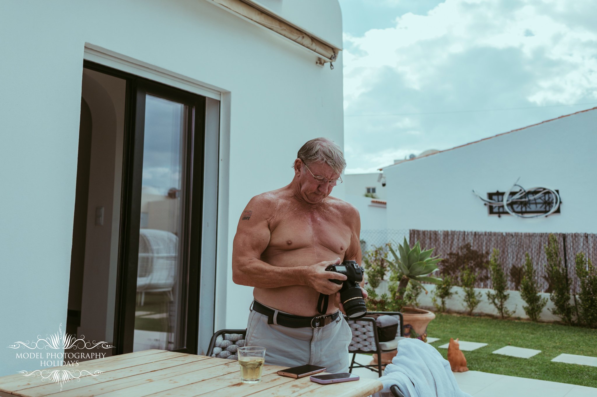 An older man with gray hair and glasses is shirtless, holding a camera and standing outside in a backyard near a patio table with a glass of drink and a smartphone on it. There is a garden with a small dog or cat and plants, a white house with slidin