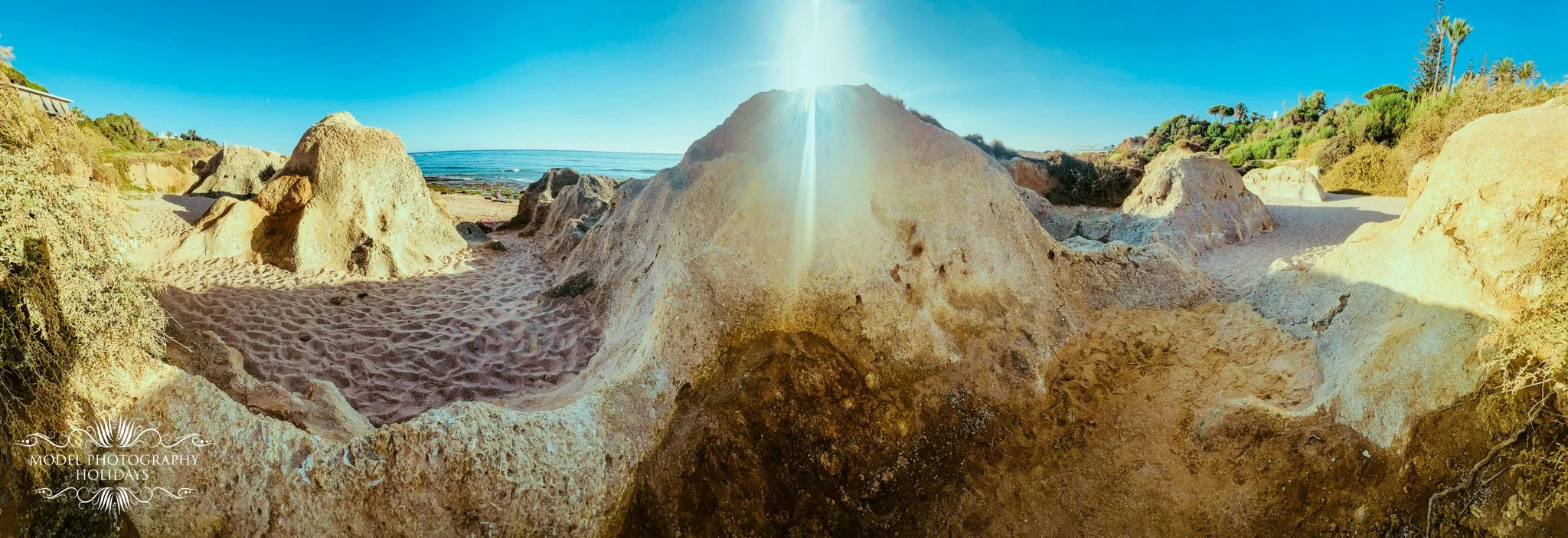 A panoramic view of a sunny beach with large rocks, sand, greenery, and the ocean in the background under a clear blue sky.