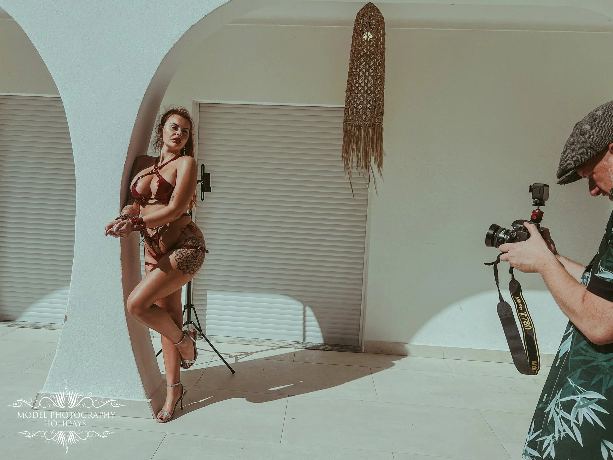 A woman posing in lingerie behind a mirrored surface with a photographer taking her picture. The scene is set indoors with white walls and rolling shutters covering windows. The woman has tattoos on her thigh and arm, and is wearing high heels and je