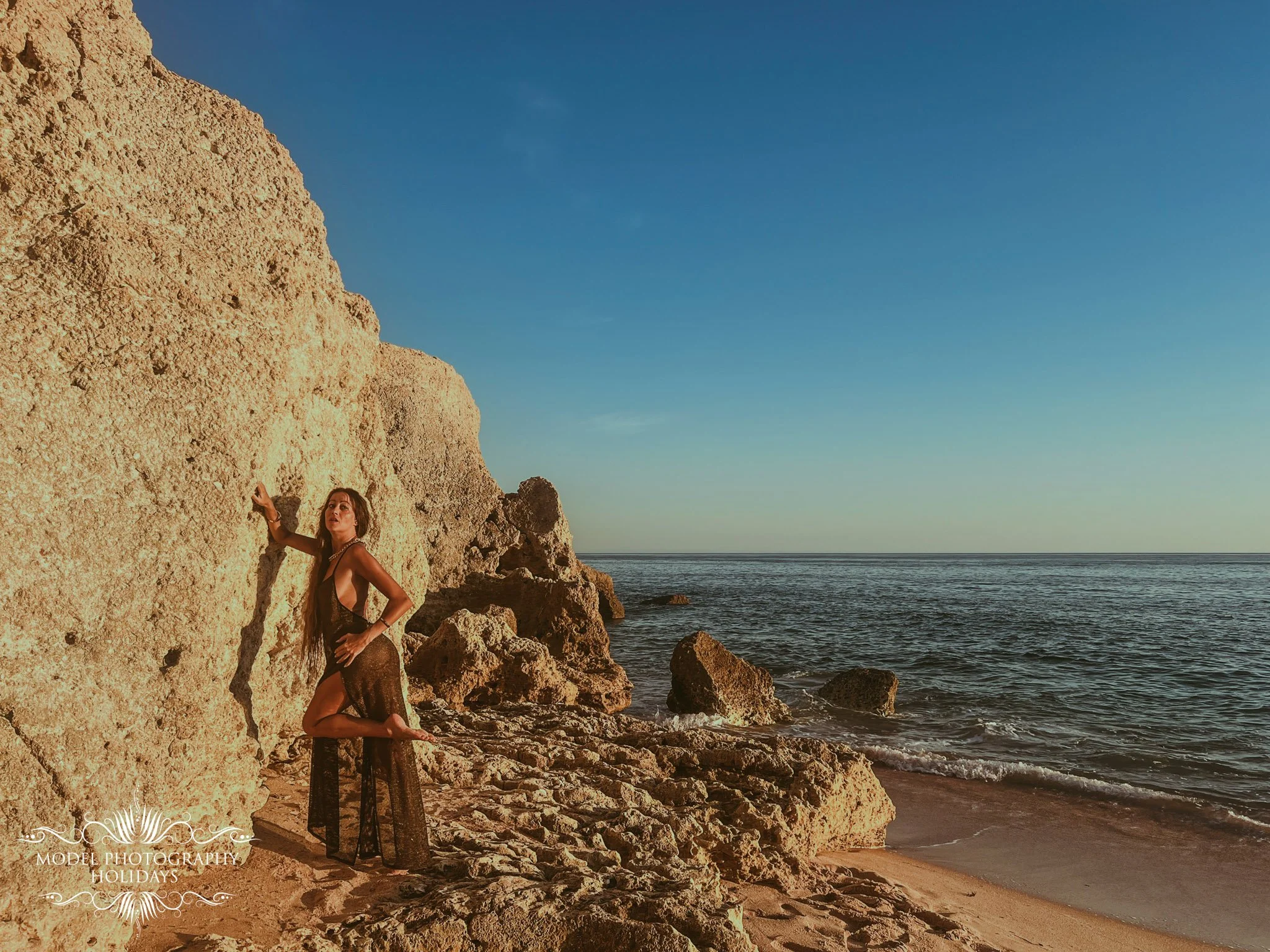 A woman in a black dress standing on rocks by the beach, with a large rock formation on her left and the ocean in the background.