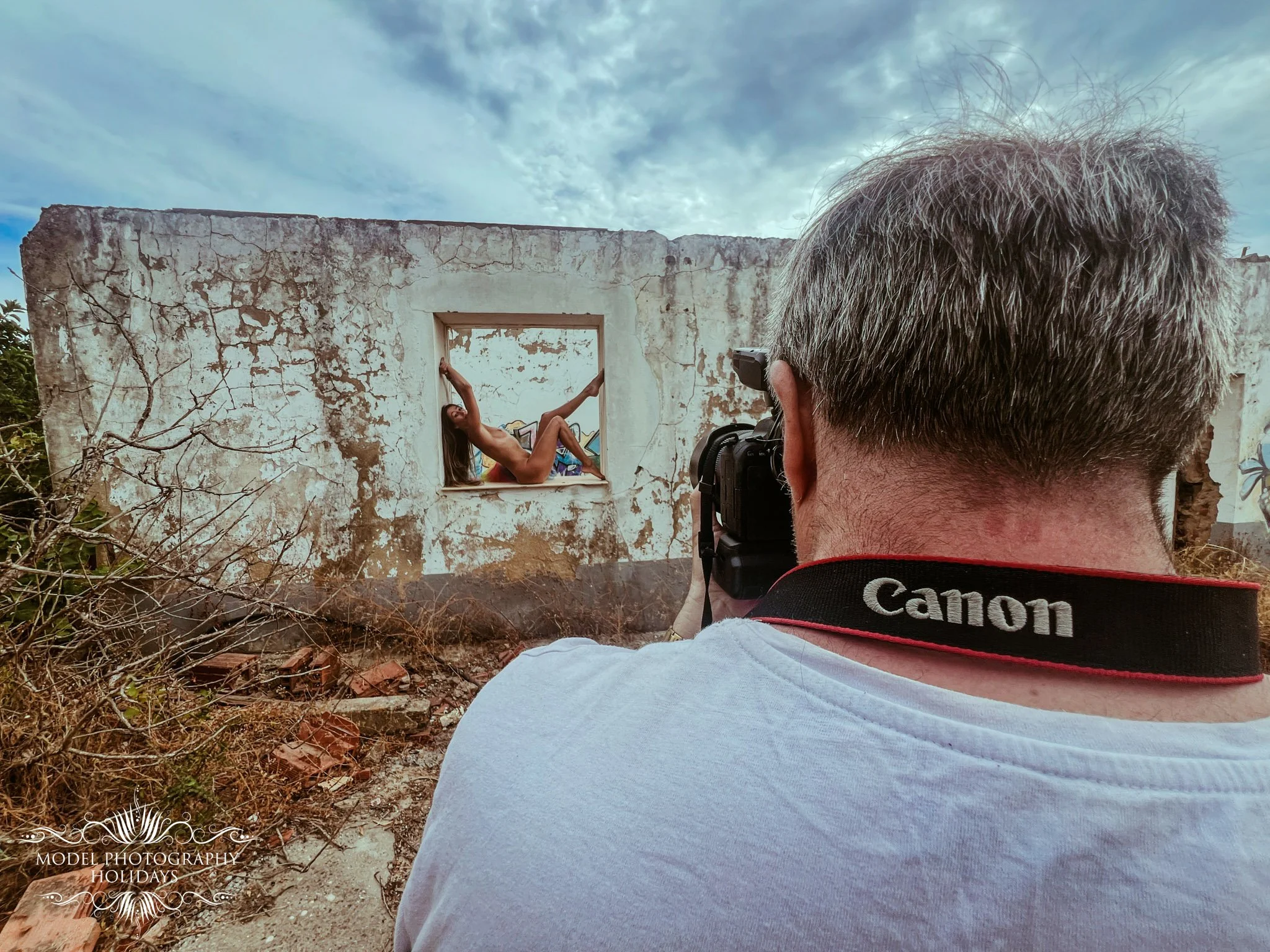 A man with gray hair taking a photograph of a woman posing in a window of a dilapidated, weathered building with cracked, peeling paint. The woman appears to be topless, and the sky is cloudy.