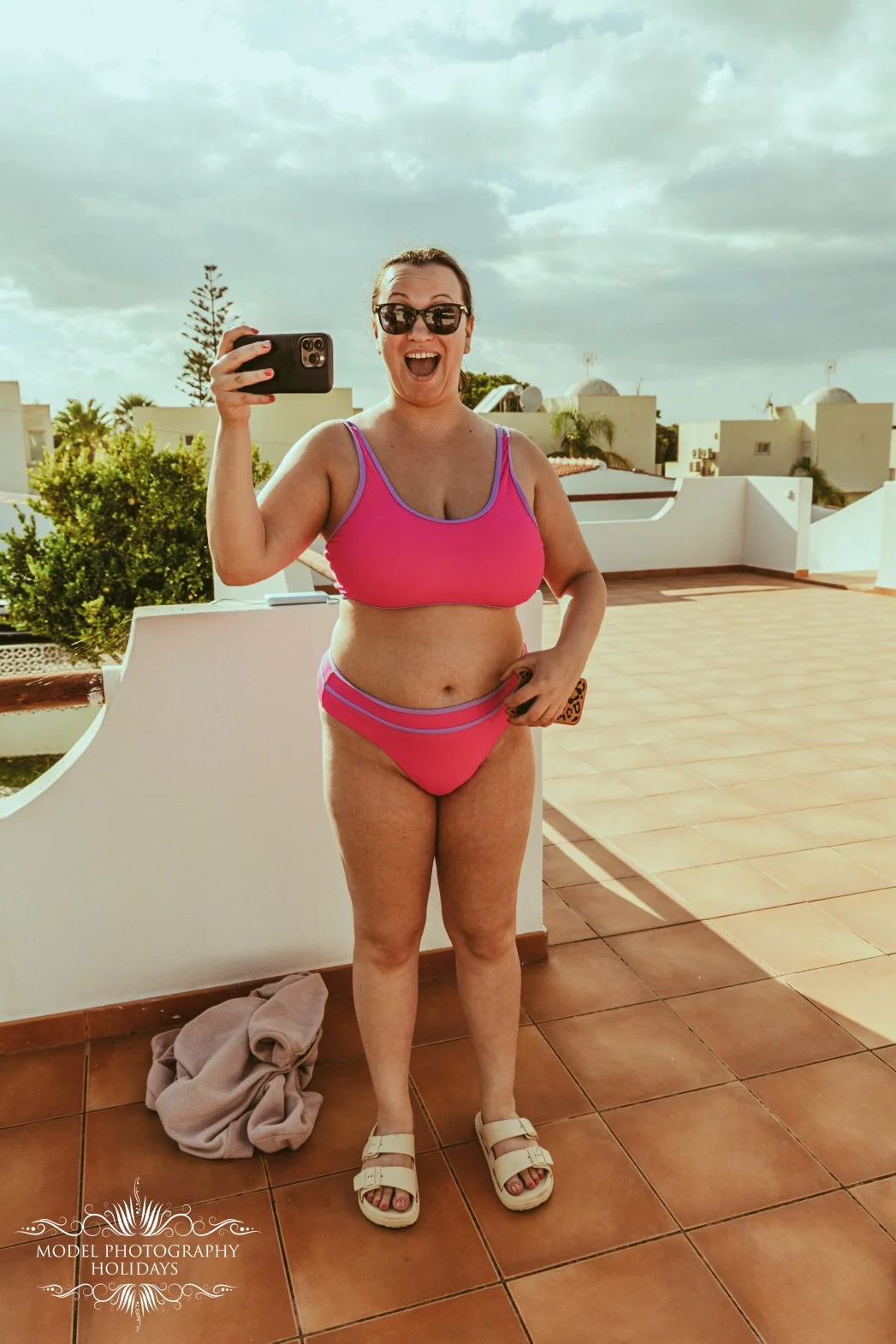A woman in sunglasses and a pink bikini taking a selfie on a terrace with a scenic background of trees, buildings, and a cloudy sky.