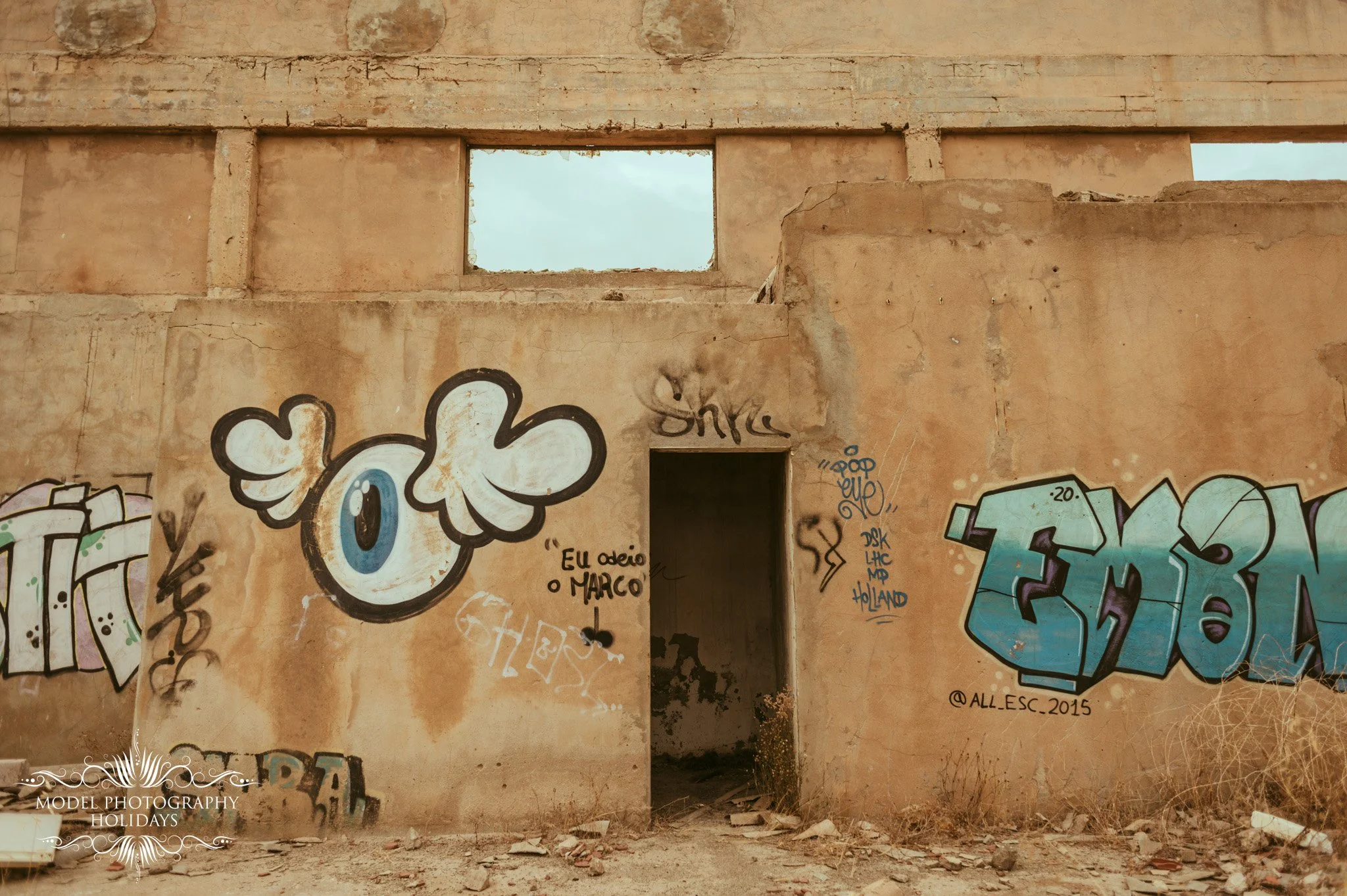 A wall with graffiti art, including a cartoon eye with a hand over it, and various colorful tags and signatures, with a cloudy sky visible through a rectangular opening in the upper part of the wall.