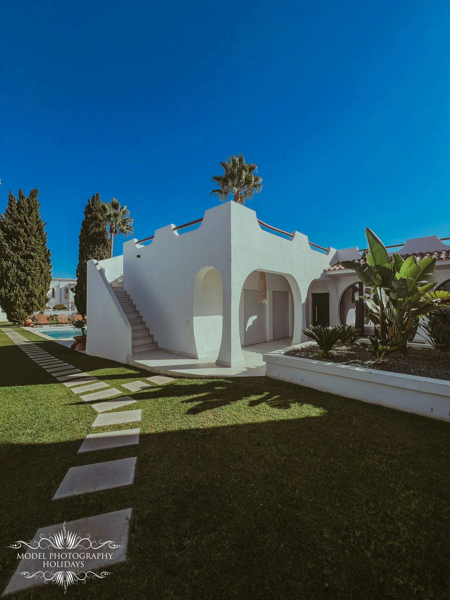White villa with steps leading to upper courtyard, palm trees, lush green lawn, and clear blue sky.