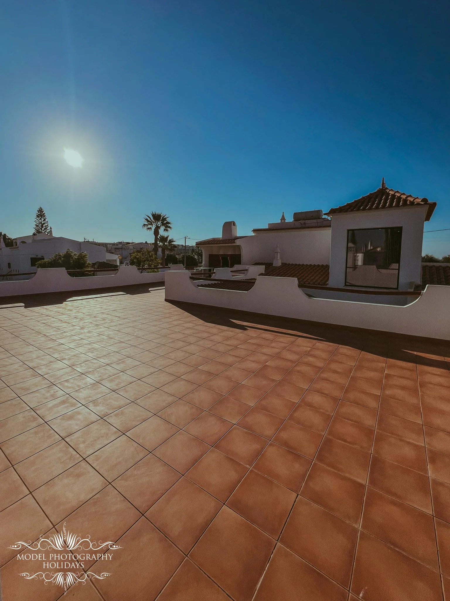 An outdoor terrace with terracotta-colored tiles, surrounded by white stucco walls, with a building featuring a tiled roof and large window, under a bright sun in a clear blue sky.