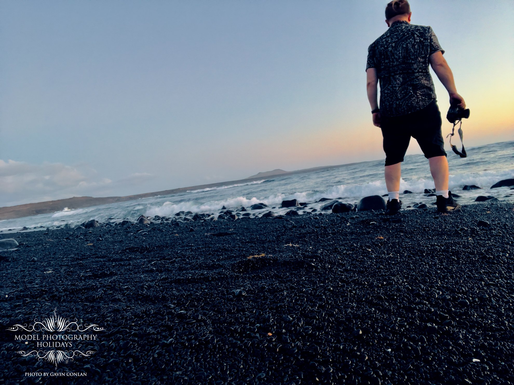Person standing on a rocky beach at sunset, holding a camera, with waves crashing in the background and cloudy sky above.