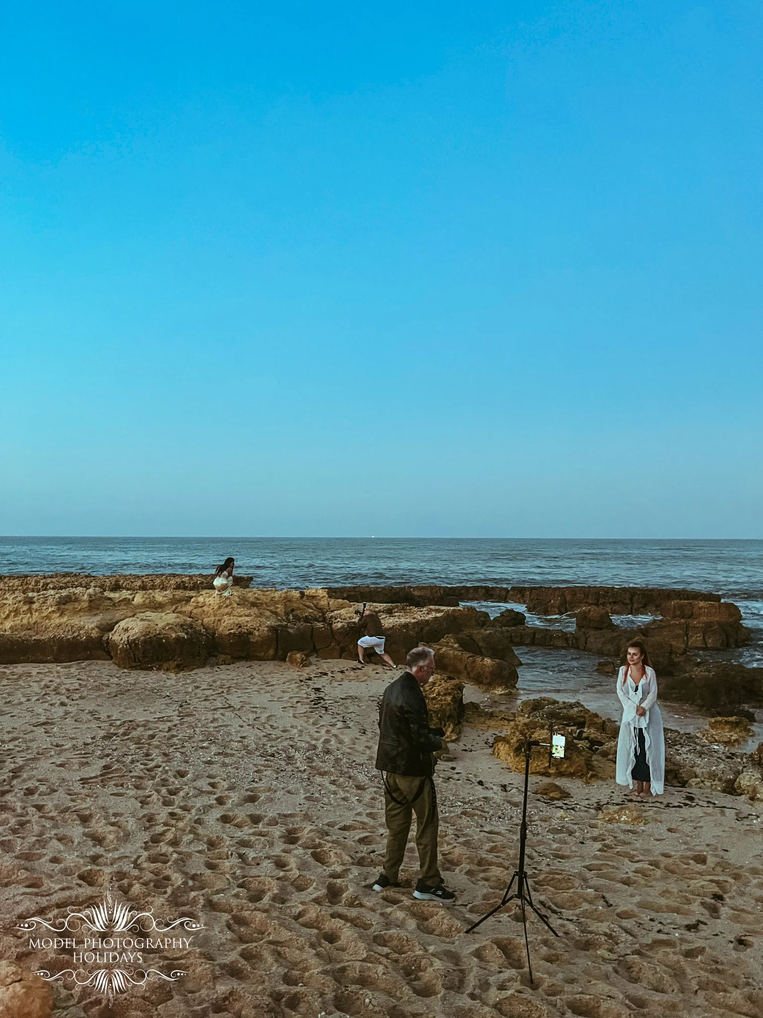 A model in a white dress is posing on a rocky beach while a photographer and an assistant take pictures. The scene is set during dusk or dawn with a clear sky and ocean in the background. There is a sign at the bottom left corner that says 'Model Pho