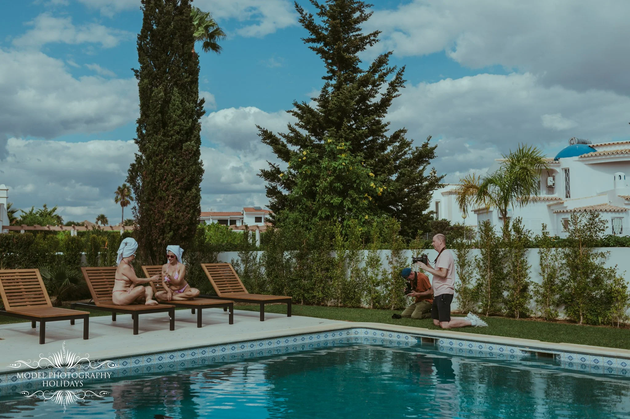 People sitting and kneeling by a swimming pool on a sunny day, with trees and white houses in the background.