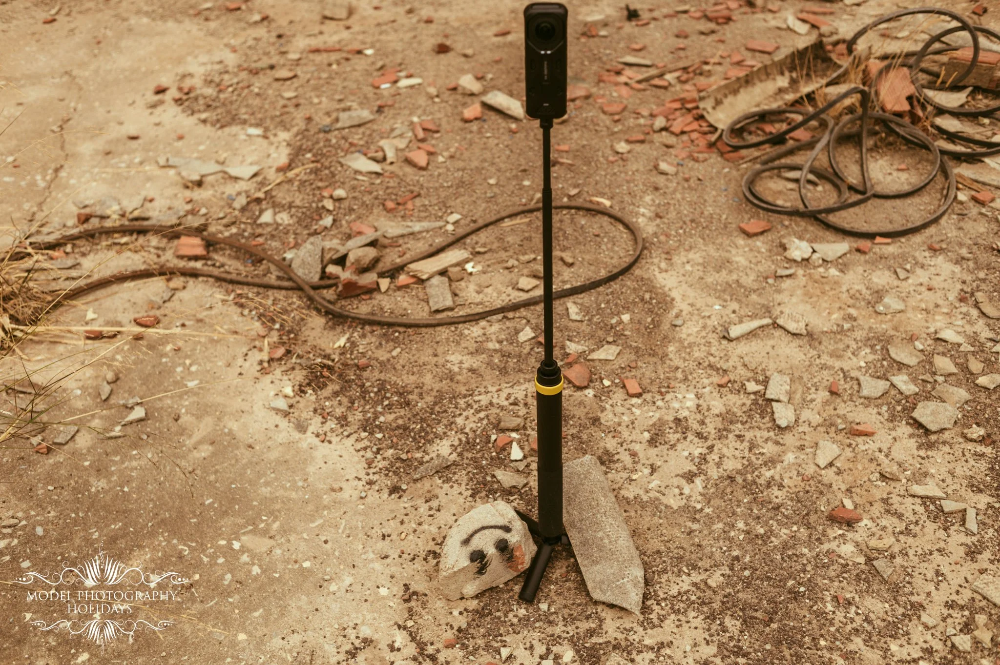 A soil surface with scattered small bricks, stones, and a pile of rubber hoses on the right side. In the center, a small black device on a yellow and black tripod stand, likely a portable scanner or sensor. The bottom left has a decorative logo with 