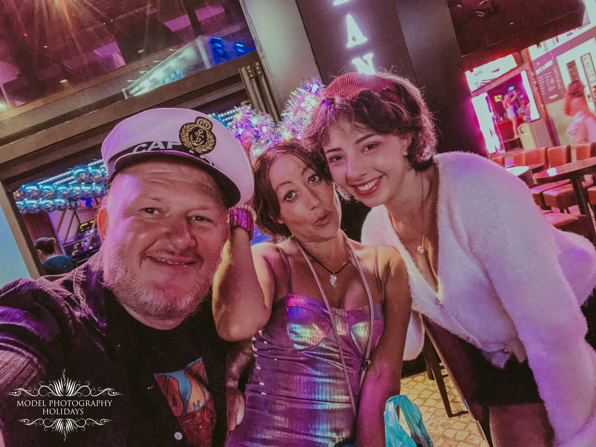 A group of three people taking a selfie together in a colorful, vibrant indoor setting with neon lights and various decorations in the background. Two women and one man are smiling and appear to be enjoying their time.