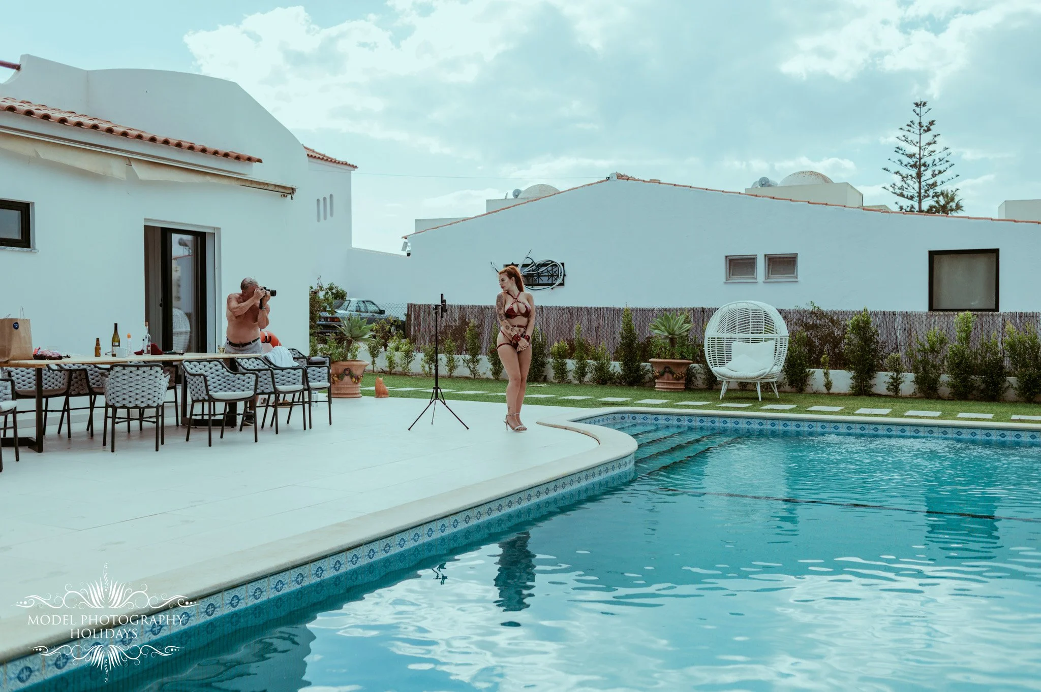 A woman in a swimsuit standing by a swimming pool while a man with a camera takes her picture. The background shows white houses with red tile roofs and some outdoor furniture, plants, and a tree.