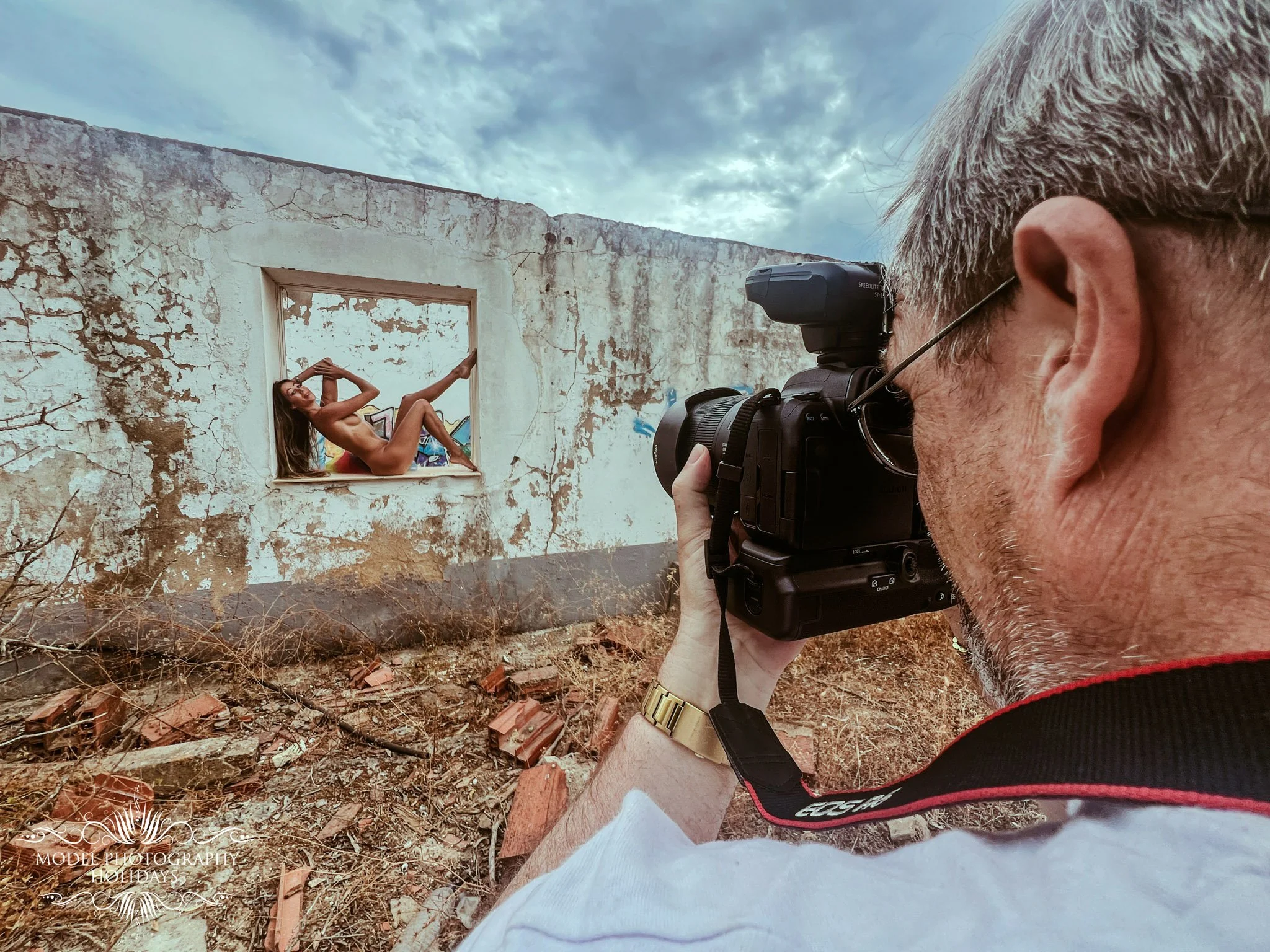 A man with gray hair and glasses takes a photo of a woman sitting in an empty window frame of an abandoned, weathered building. The woman is sitting with her back resting on the window sill, one leg bent and the other extended, with her arm behind he