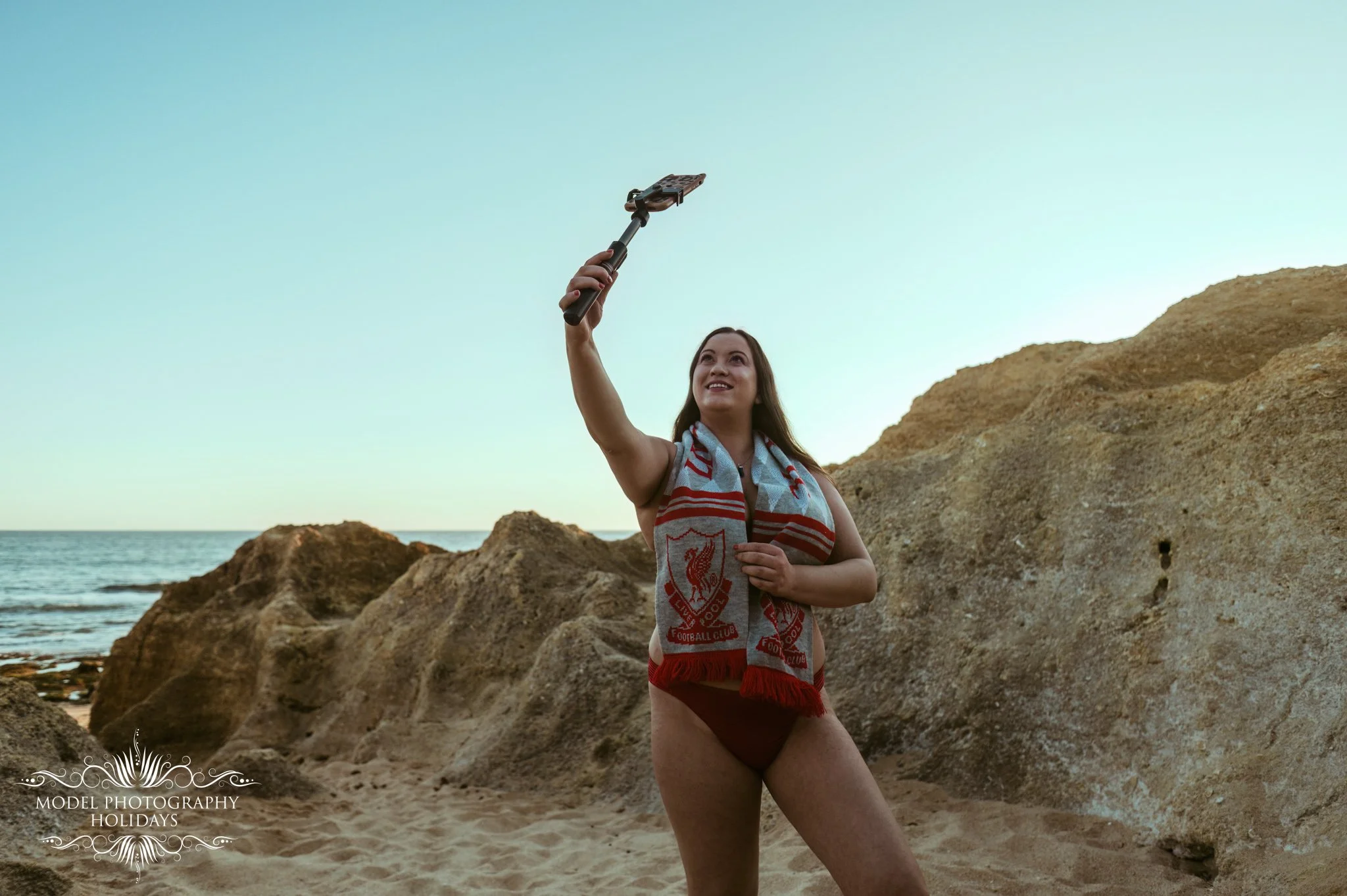 Woman in a Liverpool Football Club scarf taking a selfie on a rocky beach at sunset.