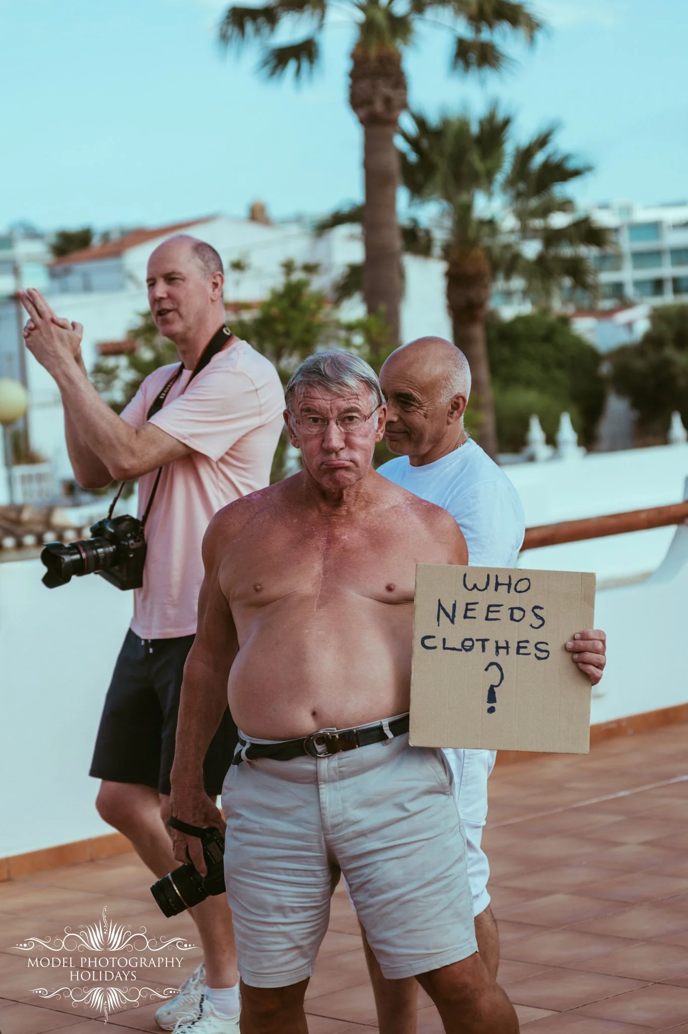 A shirtless man holding a cardboard sign that says "WHO NEEDS CLOTHES?" stands on a balcony with palm trees and buildings in the background. Two other men are behind him, one with a camera and the other smiling.