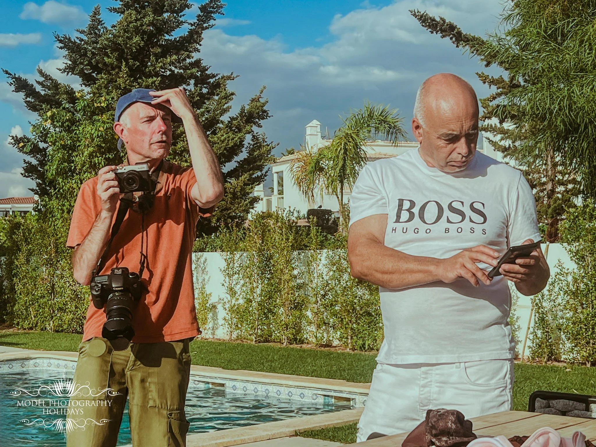 Two men outdoors near a pool, one holding a camera and wearing a blue cap, the other using a smartphone, with trees and a white building in the background.