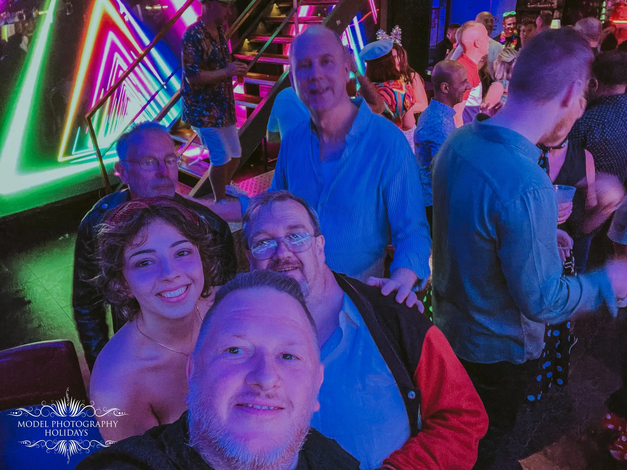 People at a party or nightclub with vibrant neon lighting, some taking selfies, and a staircase in the background. A watermark reads 'Model Photography Holidays'.