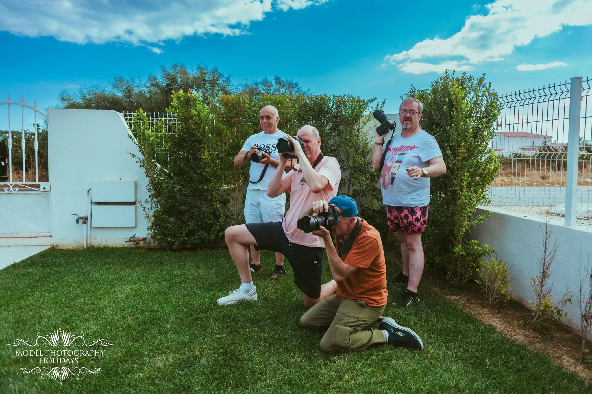 Four men taking photos outdoors in a garden, with some kneeling and others standing, under a blue sky with clouds, surrounded by green bushes and a white fence.